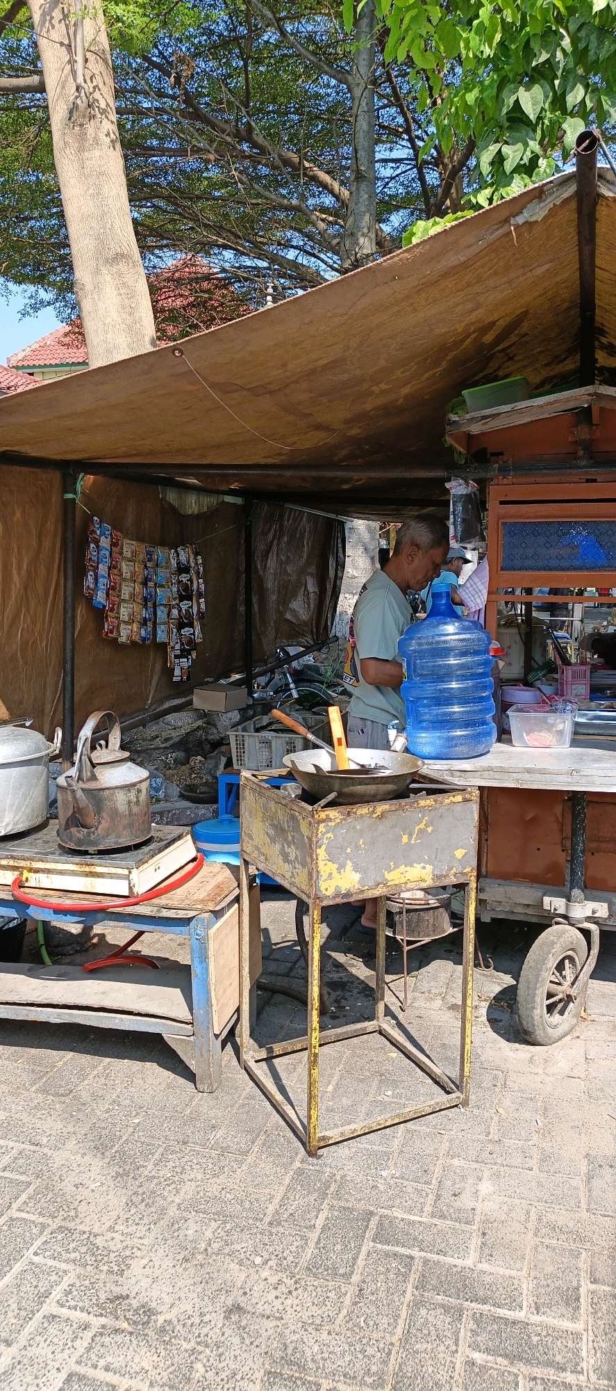 The image depicts an outdoor food stall in Klaten, Indonesia, under a makeshift tarp roof. A man, appearing to be the vendor, is shown with his head lowered, seemingly engaged in preparing food or attending to a task. In front of him is a large blue water dispenser, a wok on a metal stove, and various cooking implements. To the left, a metal kettle sits on what appears to be a hot plate. The stall is stocked with sachets of instant beverages or food items, neatly hung on display. The ground is paved with interlocking bricks. A motorcycle is visible in the background. The environment suggests a street-side operation, common in many Indonesian cities like Klaten.