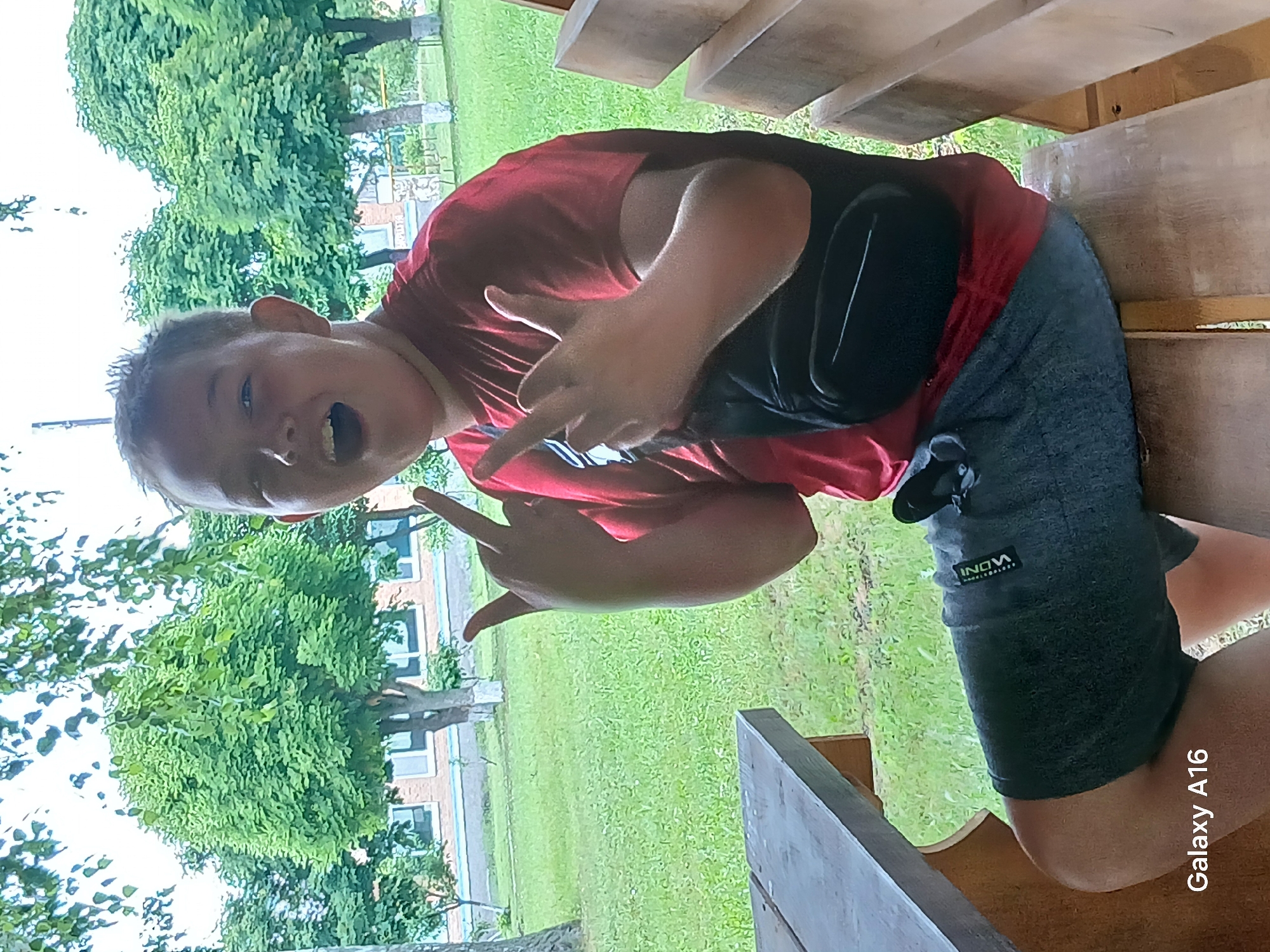 A young boy with light skin and short, dark hair is sitting on a wooden bench outdoors. He is wearing a red t-shirt and dark shorts. A black fanny pack is worn across his chest. He is making a hand gesture with both hands, extending his index and middle fingers. The boy is looking towards the camera with his mouth open. He is positioned in front of a grassy area with trees and a building in the background. The scene appears to be in Ustyvytsia, Ukraine.