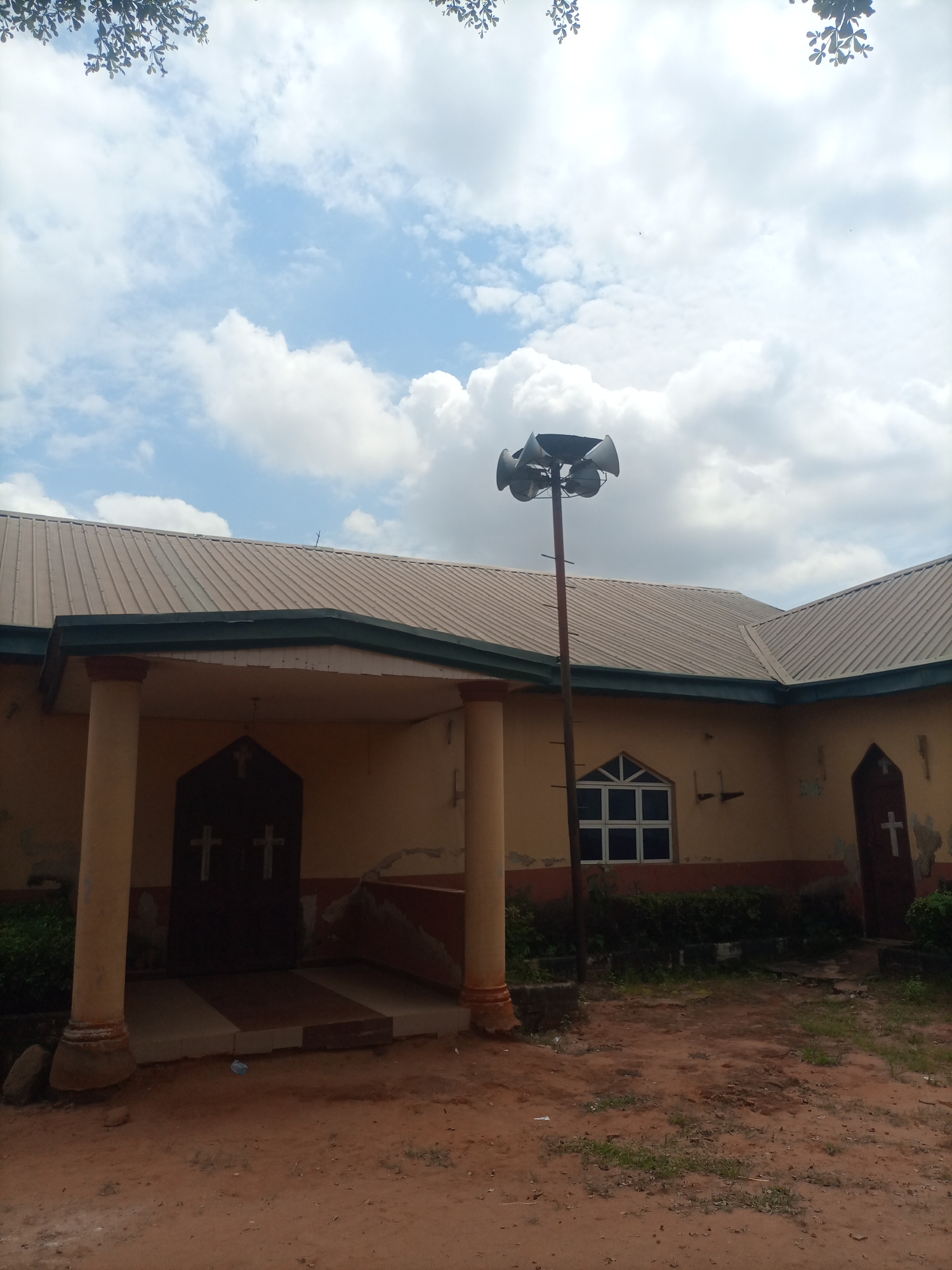 The image depicts the exterior of a church building located in Nkpor, Nigeria. The building has a metallic roof and pale yellow walls. Prominently featured is a tall pole with a cluster of multiple loudspeakers mounted at its top, positioned in front of the building. To the left, there is an entrance with two large dark doors, each adorned with two white crosses. To the right of the entrance, a white-framed window with a grid pattern is visible. Further to the right, another dark door with a single white cross is present. The ground in front of the church is covered with reddish-brown earth and sparse green vegetation. The sky above is partly cloudy with a mix of white and grey clouds against a light blue background. Tree branches with green leaves are visible at the top edge of the image.