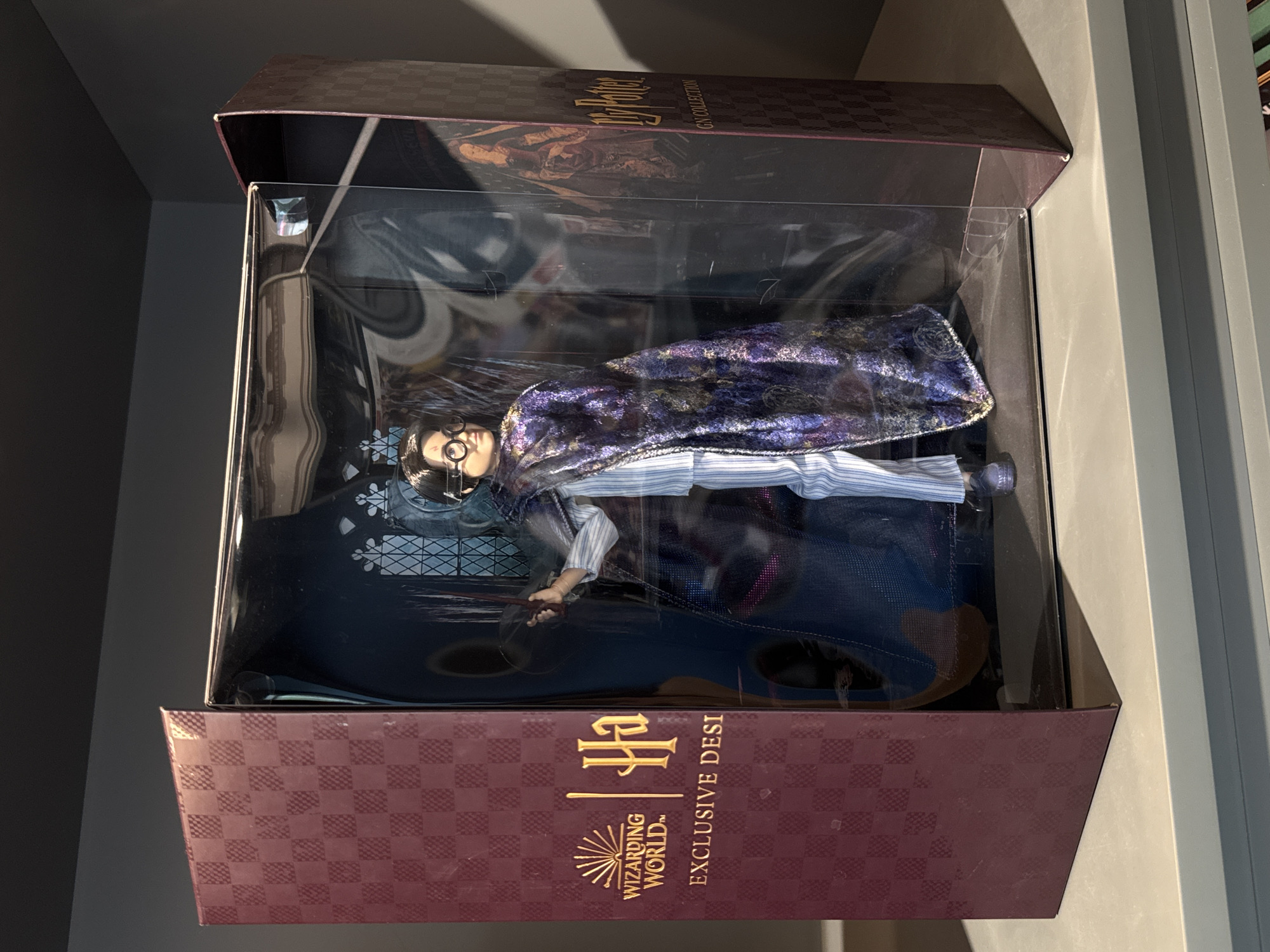 This media file features a Harry Potter doll, dressed in a blue and white striped shirt and a shimmering dark purple cloak with gold accents, lying within a clear plastic display box. The doll has dark hair and wears round glasses. The box itself is designed to resemble a book, with a checkered burgundy pattern and gold lettering on the exterior. Visible text on the box includes "Harry Potter," "GN COLLECTION," "WIZARDING WORLD," and "EXCLUSIVE DESI." The doll appears to be posed as if holding a wand. The setting is a dimly lit indoor environment, possibly a store or a personal collection display. The lighting suggests it may be daytime, with light casting shadows on the box and its contents. No specific location cues are visible beyond the implied context of a Harry Potter merchandise display. The overall impression is of a collectible item presented for viewing or sale.