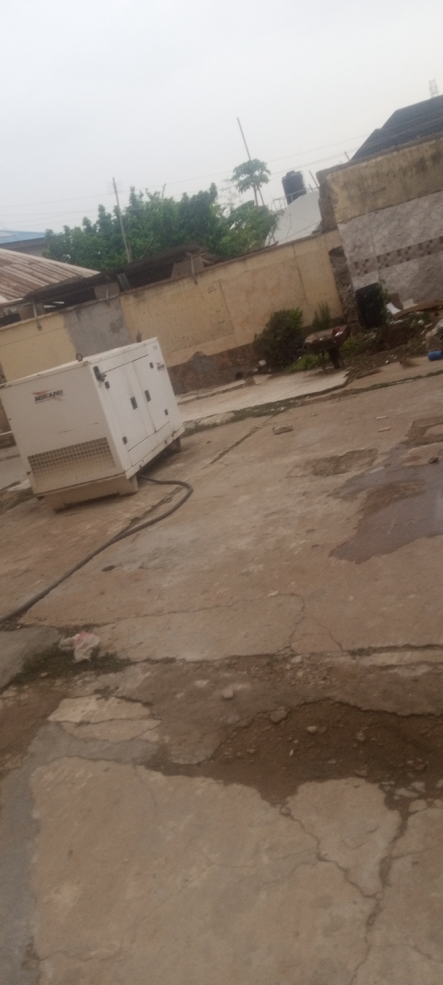 The scene shows an outdoor area in what appears to be Kaduna, Nigeria. A white, boxed generator labeled "MIKANO" sits on cracked concrete. A thick black cable runs from the generator. Behind the generator is a beige wall. Debris and overgrown foliage are present near the wall. The sky is overcast. The buildings in the background seem old.