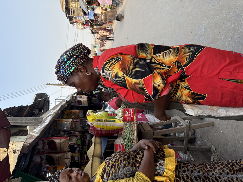 Two women at an outdoor market. One in a red floral dress and iridescent headdress leans over a vendor stall stacked with food supplies. Another woman in a yellow and brown patterned dress is reclining, eyes closed, at the bottom of the frame. Various market stalls and people are visible in the background, under a clear sky. The image is taken from a slightly elevated angle.