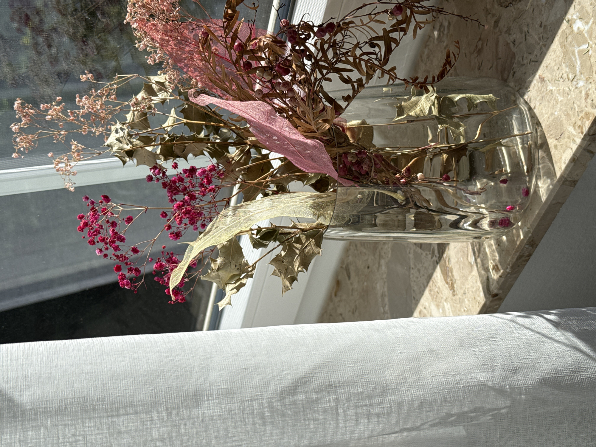 The image shows a clear glass vase filled with dried flowers and foliage, placed on a marble windowsill. A white curtain is draped on the left side of the frame, partially obscuring the window. Sunlight streams through the window, casting shadows on the windowsill and illuminating the dried floral arrangement. The flowers are a mix of delicate pink and deeper fuchsia clusters, with brown, dried leaves and stems. A few translucent, skeletal leaves, one pink and one yellowish, are interspersed among the dried elements. The background outside the window shows a blurry reflection of trees and possibly a building, suggesting an outdoor setting. The overall scene is tranquil and highlights the beauty of dried natural elements bathed in natural light. This image was captured in Bad Sachsa, Germany, specifically on the windowsill of a room facing a garden. There are no people visible in the image, and no actions are taking place.