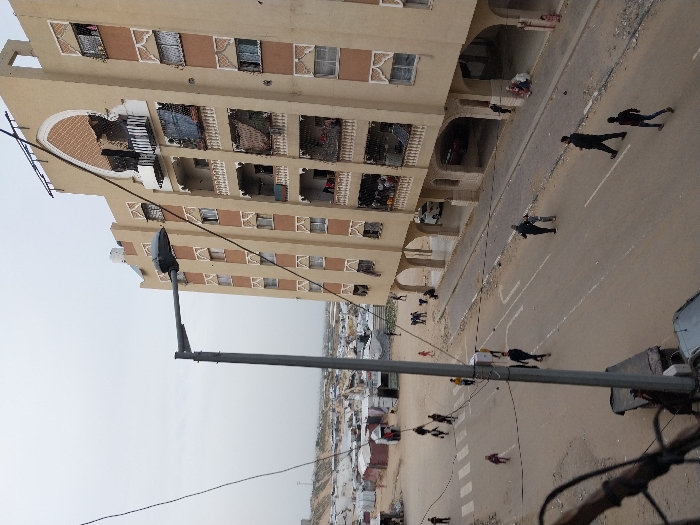 An elevated view captures a street scene in Khan Yunis, Palestinian Territory, under an overcast sky. A prominent gray street lamp post is in the mid-left, with black electrical wires crossing the upper frame. Below, a paved road with faded white pedestrian crossing stripes and a double solid line dissects the image.
To the left of the road, an expansive unpaved, sandy area transitions into a dense, sprawling encampment of numerous white and light-colored temporary shelters and tents, extending towards the horizon. Several individuals walk through this unpaved area, including a child in a red garment.
On the right side of the road stands a multi-story building, approximately seven visible floors, with a light beige facade featuring Middle Eastern architectural elements. The building has numerous balconies with white decorative balustrades; several balconies are obscured by dark tarps, blankets, or show signs of damage. Windows throughout the structure feature patterned grilles. The top floor displays a large arched decorative recess with lattice work. The ground level of the building is supported by open arches, providing a sheltered space where several people are seated or standing.
On the paved road, various individuals are seen walking. Two men are in the immediate foreground, walking right-to-left. Further up the road, a person pushes a large white rectangular object, possibly a cart or container. Other pedestrians are scattered across the road and the unpaved areas, moving in different directions.