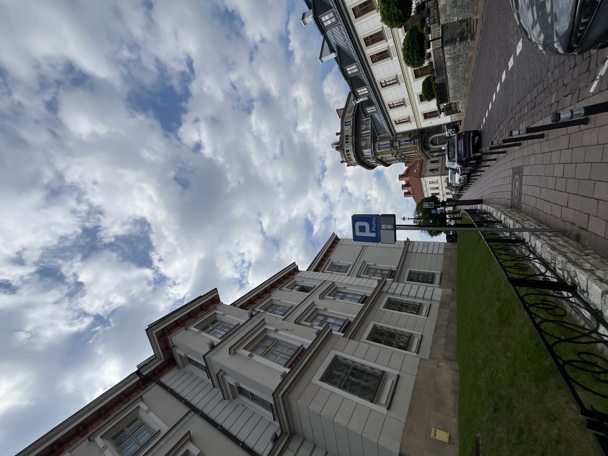 The image shows a street scene in a city, likely in Europe, with a low-angle perspective looking up towards a cloudy sky. The left side of the frame is dominated by a large, light-colored building with multiple windows and ornate architectural details. A grassy embankment slopes down from this building, bordered by a black metal fence with a decorative pattern.
To the right of the building, a paved sidewalk runs along a cobblestone street lined with parked cars. A blue and white parking sign is visible on the sidewalk. Further down the street, other buildings with various architectural styles can be seen, including a distinctive round building and a white building with a dark roof. The street appears to be relatively quiet, with only a few cars visible. The sky is filled with fluffy white clouds against a blue background. The overall impression is of an urban environment with historical architecture under a partly cloudy sky.