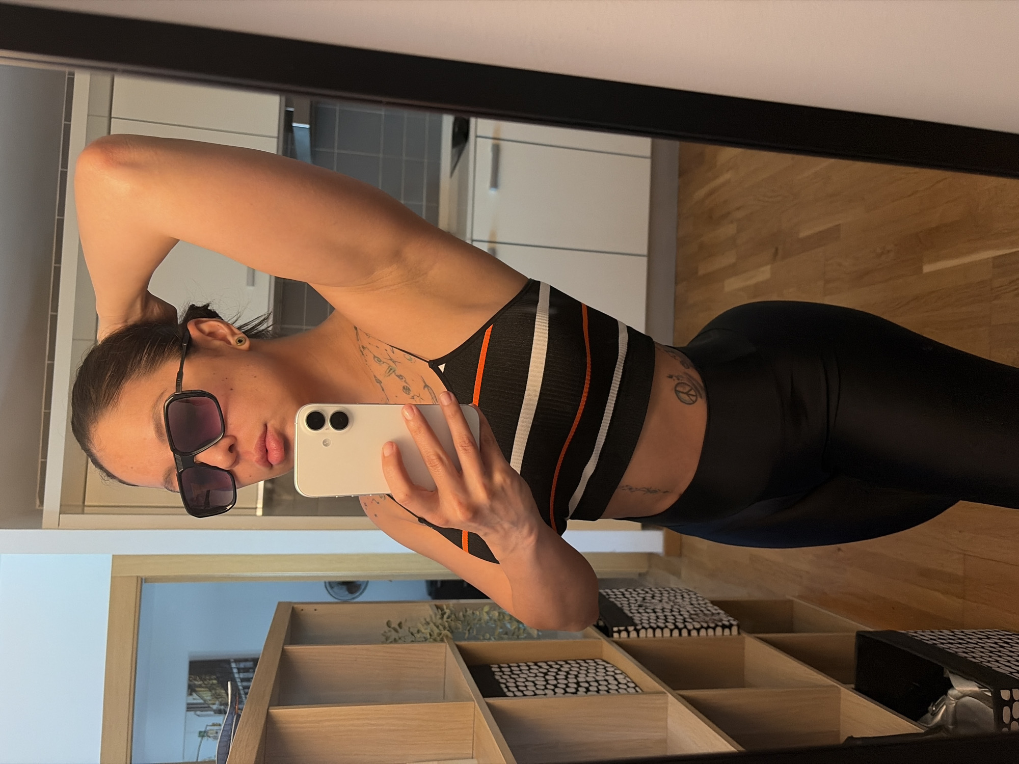 A woman is taking a selfie in front of a mirror. She is wearing sunglasses, a striped sports bra, and black leggings. Her hair is tied back. She is holding a white smartphone in her right hand. The mirror reflects a room with wooden floors, white cabinets, and a shelving unit. The location is likely in an apartment or home in L'Hospitalet de Llobregat, Spain.