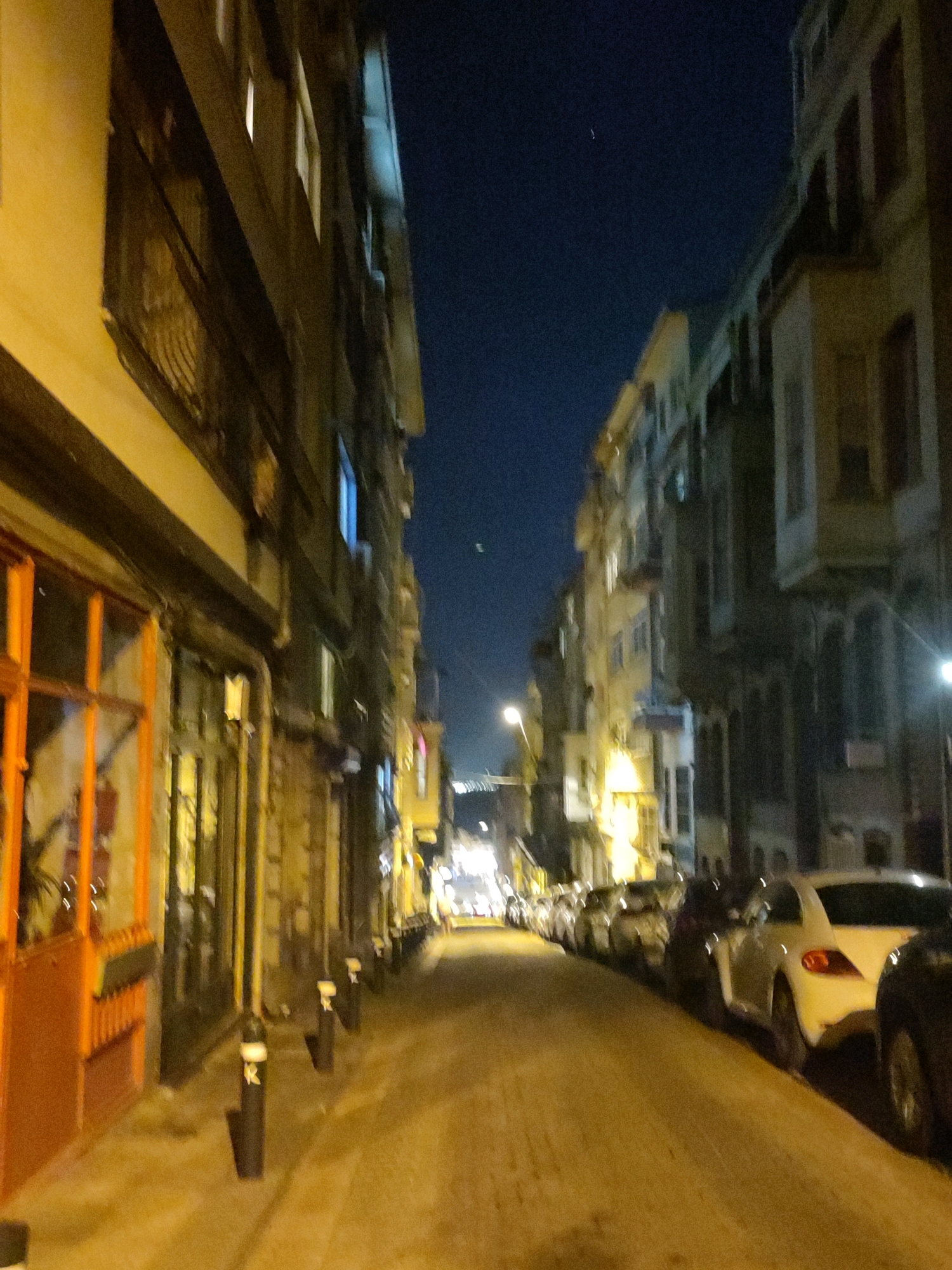 This is a nighttime photograph of a street in Kadıköy, Turkey. The street is lined with multi-story buildings on both sides, with shops or businesses on the ground floor. The buildings appear to be old and have traditional architecture. The street is illuminated by streetlights and the lights from the buildings. Several cars are parked along the side of the street. The sky is dark, indicating it is nighttime. There are no people visible in the image. The overall impression is of a quiet, urban street scene at night.