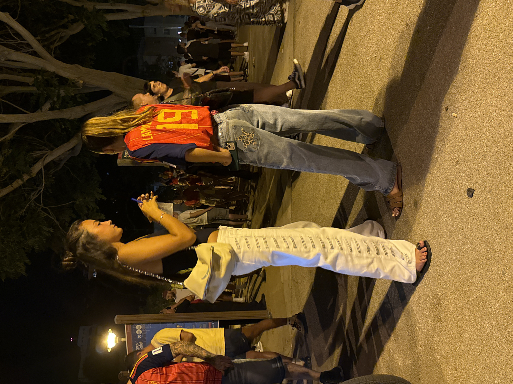 The image depicts an outdoor scene at night, likely in Blanes, Spain, with a crowd of people. Two women are the primary subjects, standing in the foreground. The woman on the left, with dark hair styled in a bun, wears a black top and loose white pants with decorative stitching. She holds a smartphone in her right hand and a white purse on her left shoulder. The woman on the right has blonde hair tied in a ponytail and is wearing a red and blue sports jersey with the number "19" and "LAMM" visible. She is also wearing light blue jeans with a distinctive leopard print and star design on the back pocket. Both women are wearing casual footwear.
The environment appears to be a public space, possibly a street or plaza, with a paved ground surface. In the background, several other individuals are visible, some appearing to be part of a gathering or event. Trees with large branches are prominent in the background, suggesting an outdoor, possibly park-like setting within the city. Lighting appears to be artificial, with a streetlamp visible on the left side of the image, casting a warm glow. The overall atmosphere suggests a lively evening event.