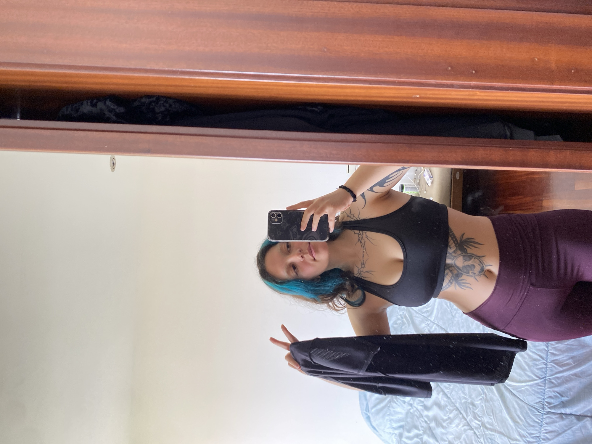 A young woman takes a selfie in front of a mirror in a room, likely her home. She is wearing a black sports bra and maroon leggings. She has blue-tinged hair and tattoos on her chest, stomach, and arm. She is holding a black jacket and making a peace sign with her fingers. The mirror reflects a bed with light blue sheets. The background includes a wooden dresser or wardrobe. It is not possible to determine whether the image was taken in Gijón/Xixón or anywhere else in Spain based on the available information.