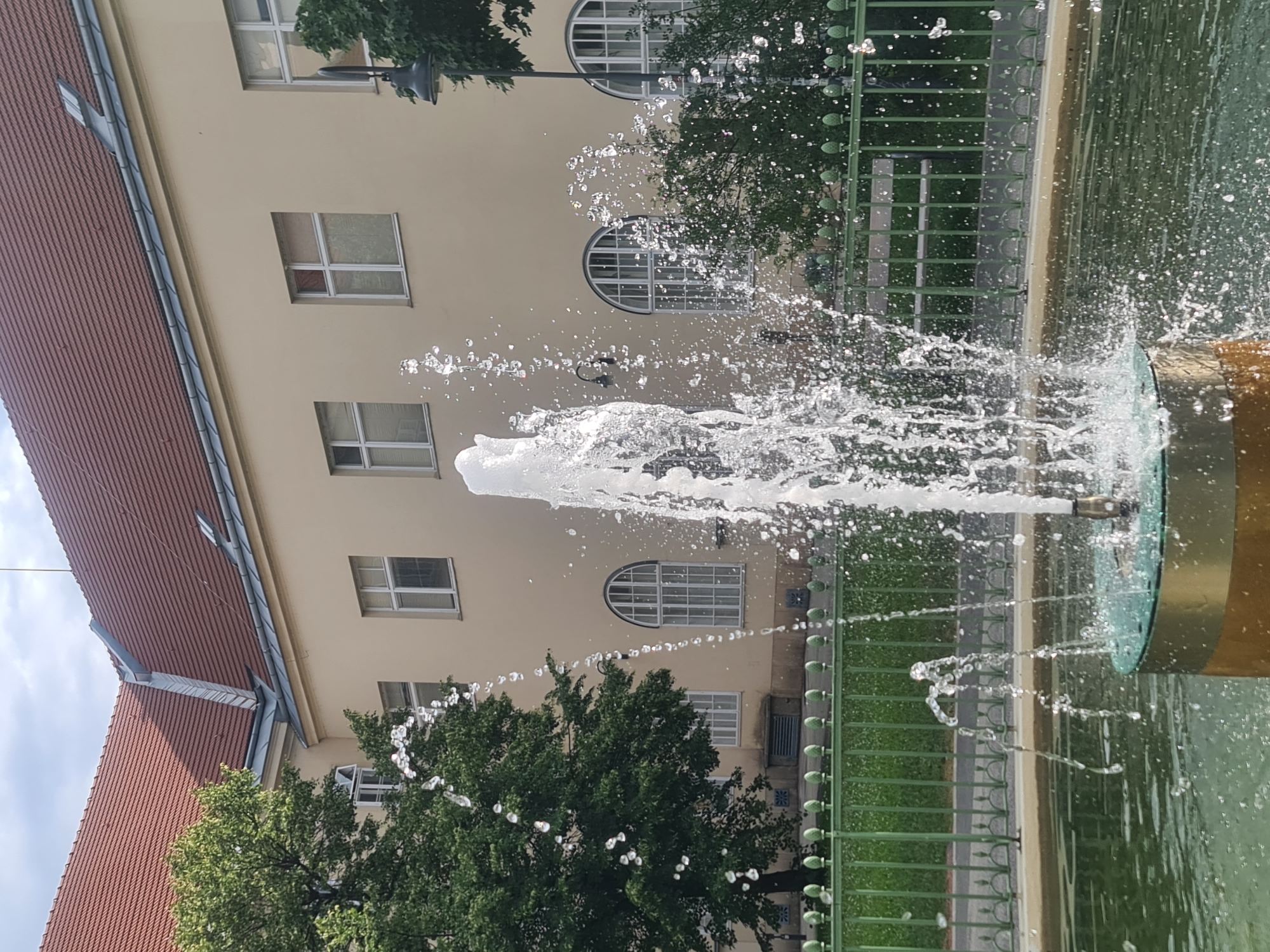 A close-up, eye-level shot captures a fountain in a courtyard, likely in Vienna, Austria, given the architectural style. The fountain, constructed from gold and metallic materials, is the primary subject, with water cascading upwards and outwards, creating a dynamic splash effect. Surrounding the fountain is a pool filled with green water. A green metal fence with decorative finials encloses the fountain area, and behind it, a park bench is visible. The background features a light-colored building with a red tiled roof and multiple windows. A large, leafy green tree stands to the left of the building, partially obscuring the fountain's left side. The sky above is overcast.