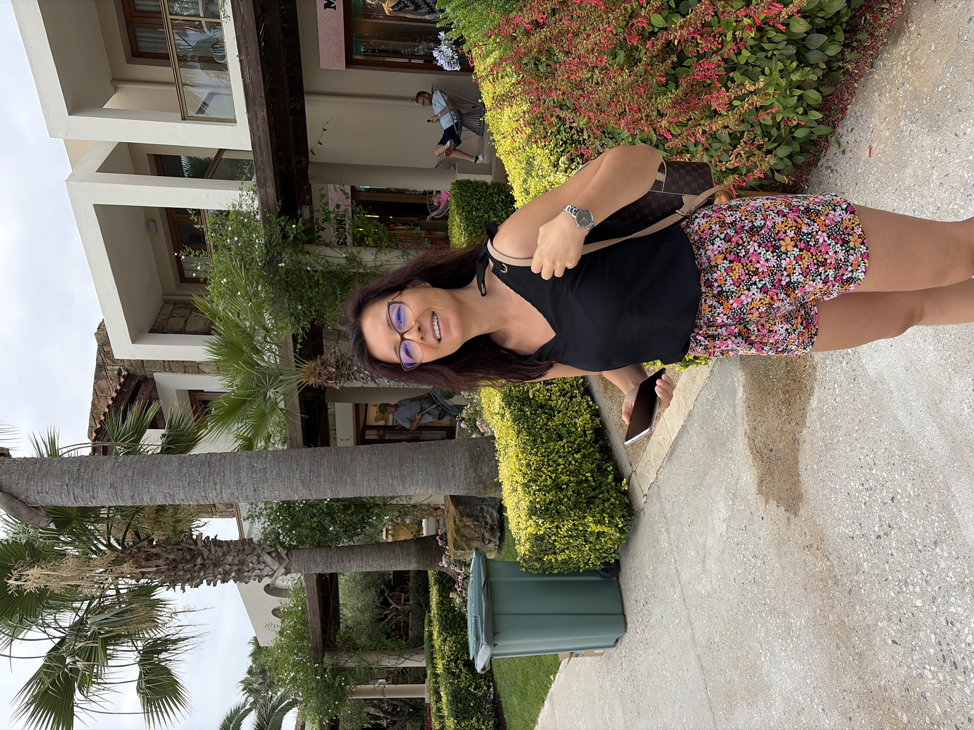 A woman with dark hair and glasses smiles while holding a phone and wearing a black tank top and floral shorts. She is standing in front of a building with shops and balconies, surrounded by palm trees and manicured bushes. In the background, a man is seen raking leaves, and another man and child are seated on chairs. A green trash can is visible to the left. The scene appears to be in a resort or hotel complex in Kassandra Municipal Unit, Greece.