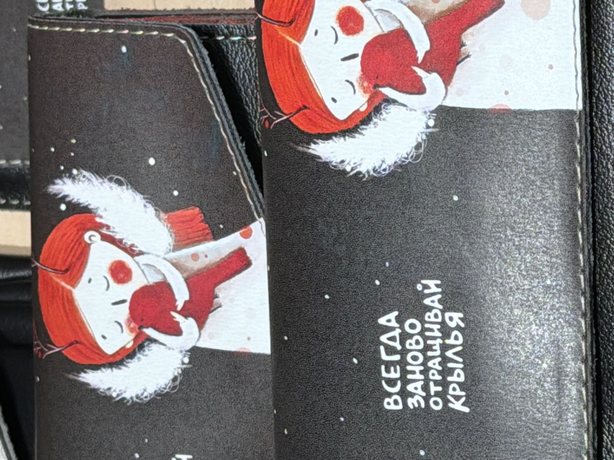 This close-up shot displays two dark, textured items, likely journals, notebooks, or wallet covers, featuring identical whimsical illustrations. Each item is adorned with an image of a stylized character, possibly a girl or an angel, with bright red hair, white skin, simple red cheeks, and closed eyes suggesting a peaceful expression. The character is dressed in red and prominently features large, fluffy white wings, giving an angelic appearance. The background of the illustration is black, speckled with small white dots that resemble falling snow or distant stars.
The items appear to be made of a textured, possibly faux leather material, with visible white stitching along their edges, indicating craftsmanship. The overall scene created by the illustration is serene and somewhat magical, evoking a winter or night-time theme.
On the right item, Cyrillic text is visible, oriented vertically and written in a playful, handwritten font. The text reads: "ВСЕГДА ЗАНОBО ОТРАЩИВАЙ КРЫЛЬЯ." This translates to "ALWAYS GROW YOUR WINGS ANEW" or "ALWAYS REGROW YOUR WINGS," conveying a message of resilience and renewal.
Given the context of Saint Petersburg, Russia, these items might be locally produced goods or souvenirs.