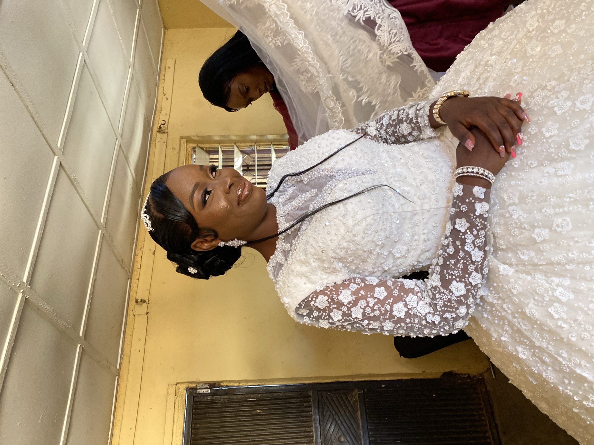 A woman in a white wedding gown stands indoors with another woman nearby. The bride has dark skin, black hair pulled back, a tiara, jewelry, and long sleeves adorned with white embellishments. Her hands are clasped. Another woman with black hair stands behind her with a white lace garment draped over her head and a maroon-colored fabric visible underneath. The setting appears to be a simple room with a pale yellow wall, a window with blinds, and a dark door. Based on the image metadata, this photograph was taken in Vom, Nigeria.