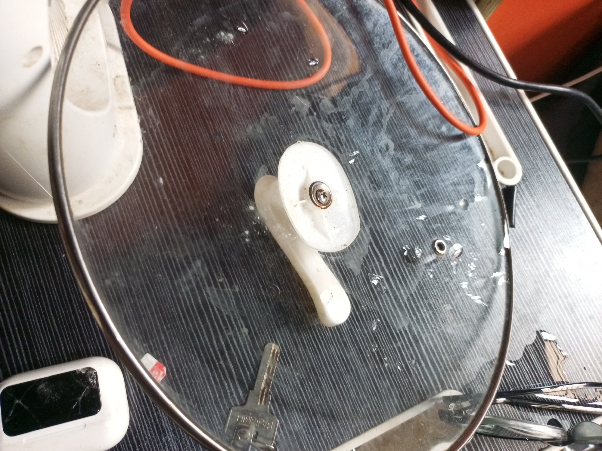 This image depicts a close-up, overhead view of a glass pot lid resting on a dark, textured surface that appears to be a table. The lid is slightly damp with scattered water droplets. A white pot lid handle, affixed with a screw, is the central object on the lid. To the left of the handle, a white object, possibly a charging case for earbuds, is visible with a cracked screen. A silver key is lying on the surface to the lower left of the handle. A bright orange electrical cord and a black electrical cord are also present on the lid, suggesting they are part of a nearby appliance. The overall impression is of a domestic setting, potentially a kitchen counter or dining table. The location is specified as Ikorodu, Nigeria.