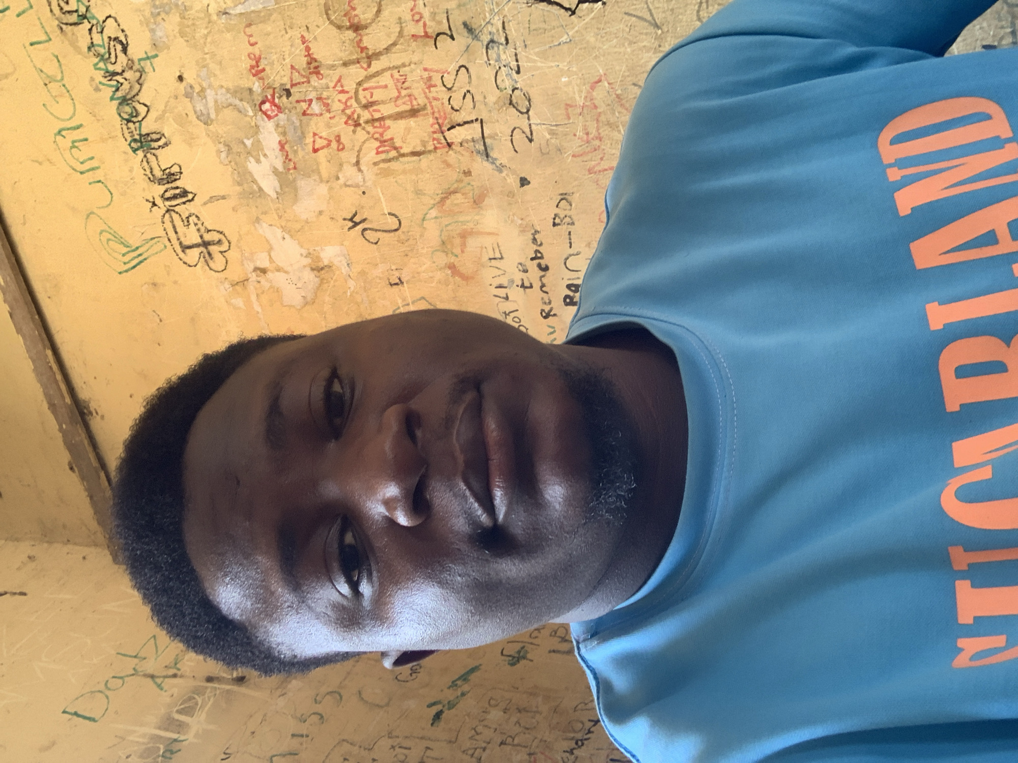 A man with dark skin and short black hair is taking a selfie in front of a wall covered in graffiti. He is wearing a bright blue t-shirt with orange lettering that reads "SUGARLAND". The wall behind him is yellow and covered in various writings and drawings in different colors, including green, black, and red. Some of the visible text includes "Dat", "PURGELL", "FIFTY", "2k", "LIVE to Remember", "Rain-Boi", and "2022". The lighting suggests an indoor setting, possibly a room or an outdoor sheltered area. The overall impression is casual and informal, with the graffiti suggesting a public or communal space.