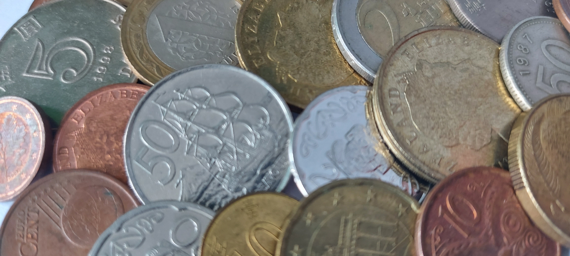 The image displays a close-up, overhead view of a collection of various coins, predominantly metallic and circular. The coins are overlapped and arranged in a disorganized manner. Some coins are silver-toned, others are gold-toned, and some are copper-toned. Visible markings on the coins include numbers, inscriptions, and designs. For example, one coin clearly shows the number "5" and the year "1998". Another coin has the inscription "ELIZABETH R" and a portrait of a person. Some coins have intricate patterns, while others are simpler. The overall impression is of a jumbled pile of currency. There is no indication or evidence within the image to suggest the location of this scene in the city of Chita, Russia, nor are there any people, actions, or interactions depicted. The environment appears to be a flat surface, likely a table or countertop, upon which the coins are placed.