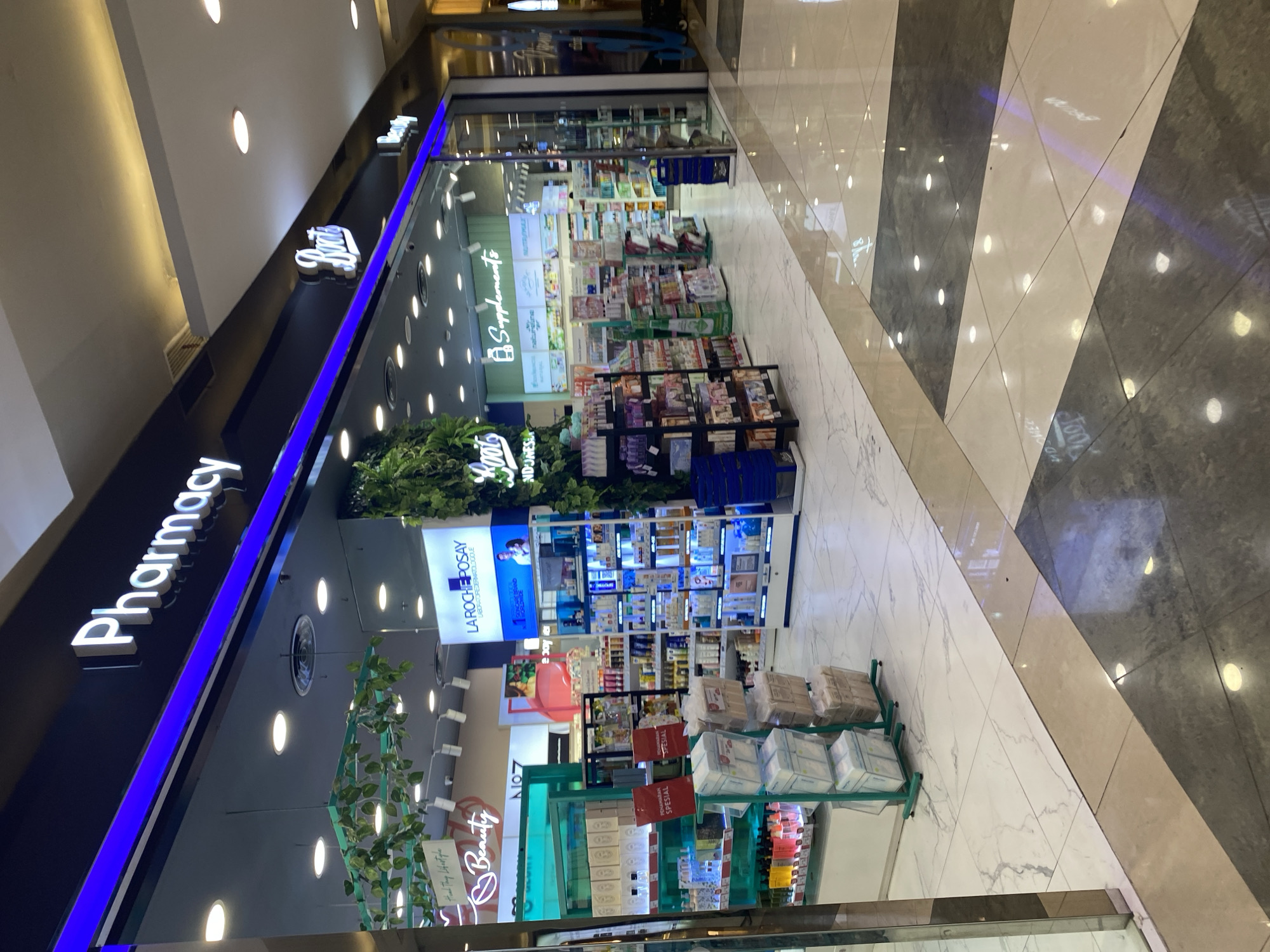 A retail store interior, likely a pharmacy or a beauty and health store, is depicted in this image. The storefront signage prominently displays "Pharmacy" in large white letters, with a blue illuminated strip below. To the right, a sign for "Boots" is also visible. The store is well-lit with overhead lights. Inside, shelves are stocked with various products, including skincare items, supplements, and other health and beauty products. A section of the store appears to be dedicated to "La Roche-Posay" products. Decorative elements like artificial plants and greenery are incorporated into the store's design. The flooring is a marble-patterned tile with geometric divisions of light and dark colors. The overall impression is of a clean, organized, and modern retail environment. The location is West Jakarta, Indonesia.