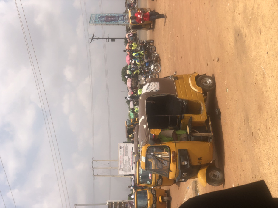 The scene depicts an outdoor transportation and commercial area in Lafia, Nigeria. The ground is unpaved, consisting of reddish-brown soil. The sky is partly cloudy with visible power lines and utility poles.
In the left foreground, a yellow three-wheeled motorized rickshaw (keke napep) with a brown fabric canopy is stationary, facing forward and slightly left. The letters "LFA" are visible on its front fender. A male driver, wearing a light-colored shirt and a green safety vest, is seated inside. A small, unadorned wooden tray is affixed to the exterior of the vehicle's left side. To the right of the foreground rickshaw, a male individual wearing a red shirt with the number "15" emblazoned on the front walks from right to left across the mid-ground.
In the mid-ground and background, multiple additional yellow three-wheeled rickshaws are parked. To the right, a cluster of motorcycles is stationary, accompanied by several individuals, many of whom are wearing bright green safety vests. Colorful market umbrellas (pink, green, blue) are visible in the background, indicating market stalls or shaded gathering points. A white rectangular sign board with unreadable text is mounted on poles in the left-center background. On the far right, a tall, blue-themed billboard displays a stylized image of a person's head. Numerous other people are dispersed throughout the background, appearing to be engaged in various activities, consistent with a transport hub or market.