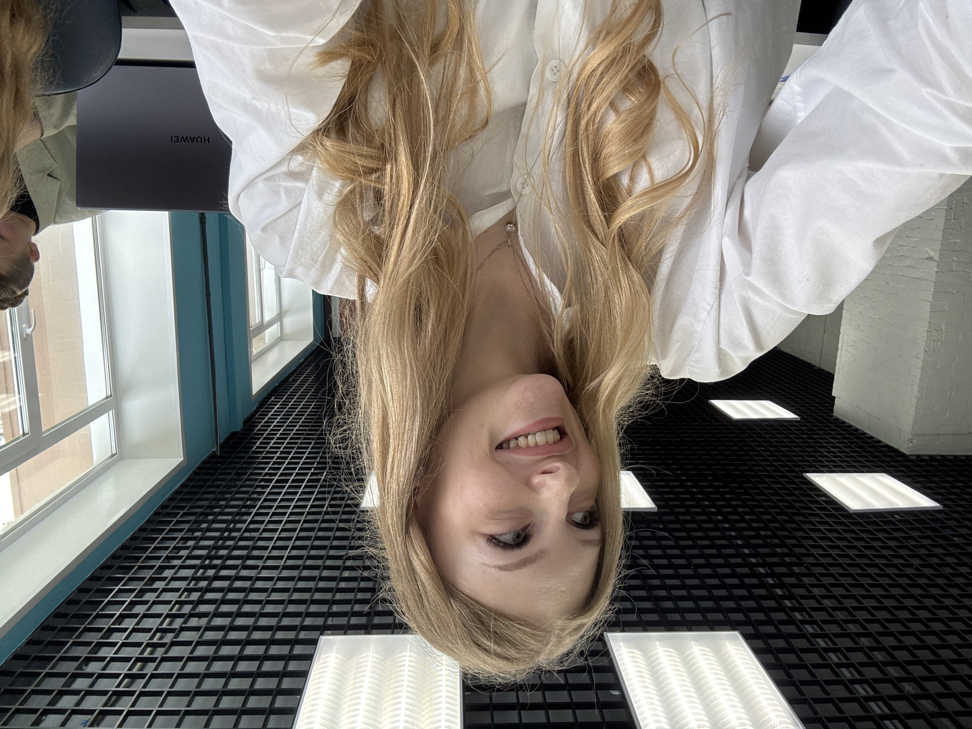 A young blonde woman is pictured in a selfie, looking off to her left with a smile. She is wearing a white collared shirt and a delicate necklace. Her hair is long and wavy, falling around her shoulders. The background shows a modern office or classroom environment with a black grid ceiling, fluorescent lighting, and large windows. A laptop with the HUAWEI logo is visible on a desk to her right. The scene appears to be an indoor space, likely an educational institution or a contemporary workspace, which aligns with locations found in the city of Perm, Russia.