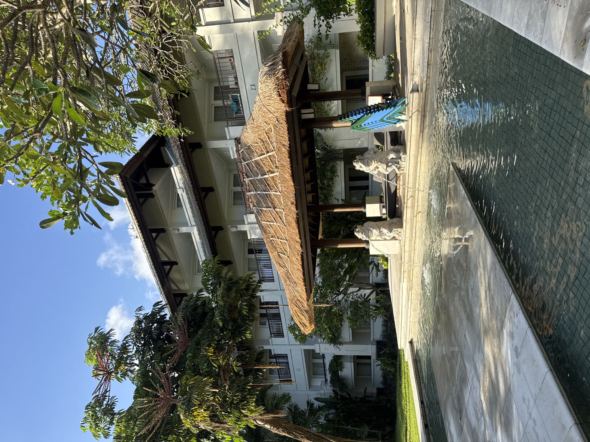 The image depicts a building in Jimbaran, Indonesia. The structure is multi-storied and white, with dark accents. A thatched-roof shelter stands adjacent to the building, supported by wooden columns. The shelter appears to house stacked chairs. To the right of the shelter is a decorative water feature with a textured surface. Lush greenery surrounds the building, including trees with visible red flowers. A clear blue sky is visible above, partly obscured by the foliage. Sculptures of animals are present on the right side of the shelter.