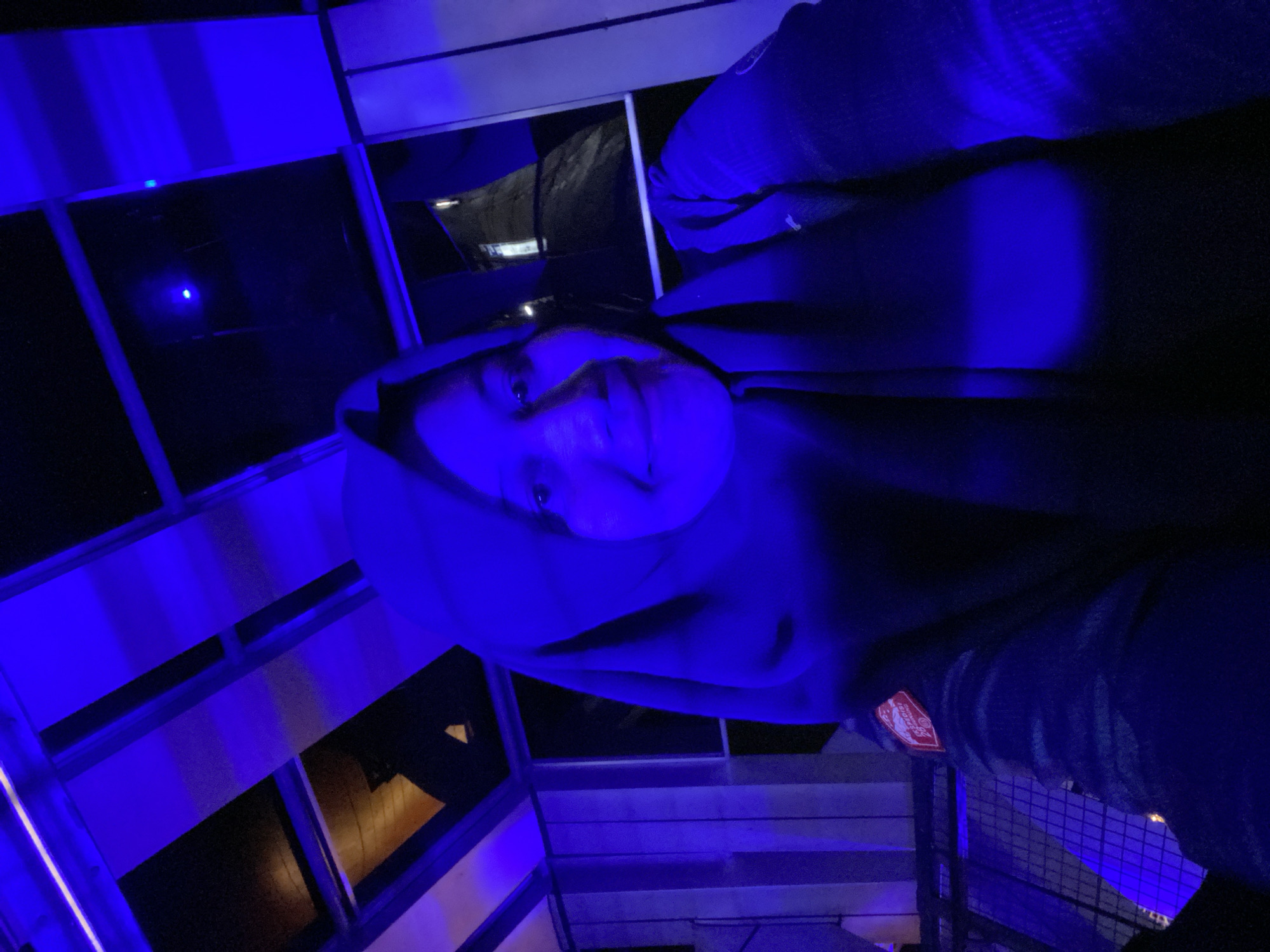 A young woman wearing a hijab and a dark jacket is captured in a selfie under blue lighting. The background reveals a structured ceiling with translucent panels and dark frames, suggesting an indoor or covered outdoor space. Vertical elements, possibly window blinds or architectural features, are visible on the left. A patch with text and an emblem is present on the woman's jacket sleeve. The lighting casts a strong blue hue across the scene, obscuring some details but highlighting the woman's face and the textures of her clothing. The location is identified as Banjarbaru, Indonesia.