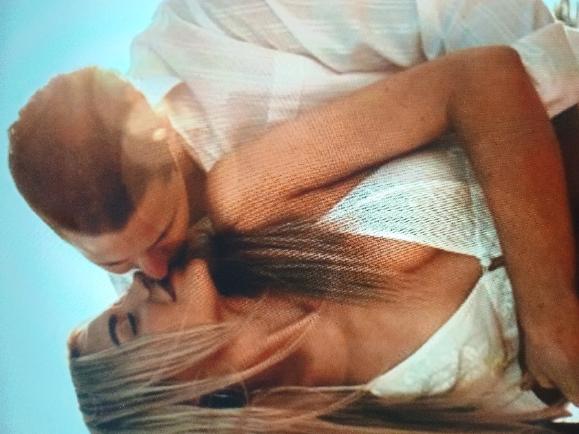 A close-up, softly focused, outdoor image depicts two individuals, a man and a woman, in an intimate embrace against a clear, light blue sky in Unknown, Unknown. The woman, with long blonde hair, has her eyes closed and lips slightly parted as she leans towards the man. She is wearing a white or light-colored textured top, possibly a bra or bikini, revealing a portion of her chest. Her left arm is visible, curving around the man's back. The man, with short dark hair, is positioned to her right, leaning in towards her. He is wearing a white, vertically textured button-down shirt. His right arm appears to be around the woman's back. Their faces are very close, with their lips appearing to be in contact or immediately preceding a kiss, indicating a romantic interaction. Bright, natural light illuminates the scene, creating highlights on their skin and clothing, suggesting a sunny day. The background is uniformly light blue and out of focus.