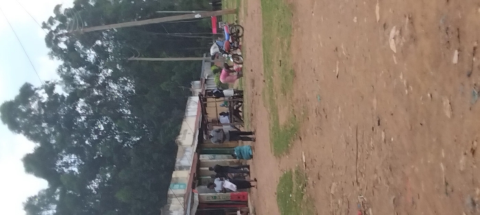 The image shows a dirt road in front of several buildings, presumably shops, in the town of Lwandeti, Kenya, located at latitude: 0.563, longitude: 34.807. There are several people present, including three men standing by the shops, two women in pink clothing standing on the road, a man and a woman standing near a motorcycle, and a woman in a blue dress sitting on the ground. There is a motorcycle parked on the side of the road, and a few other objects such as bags and chairs can be seen scattered about. The image is captured from a high angle, looking down at the scene. The buildings are made of concrete and have a corrugated metal roof. The scene is set against a backdrop of dense, green trees.
