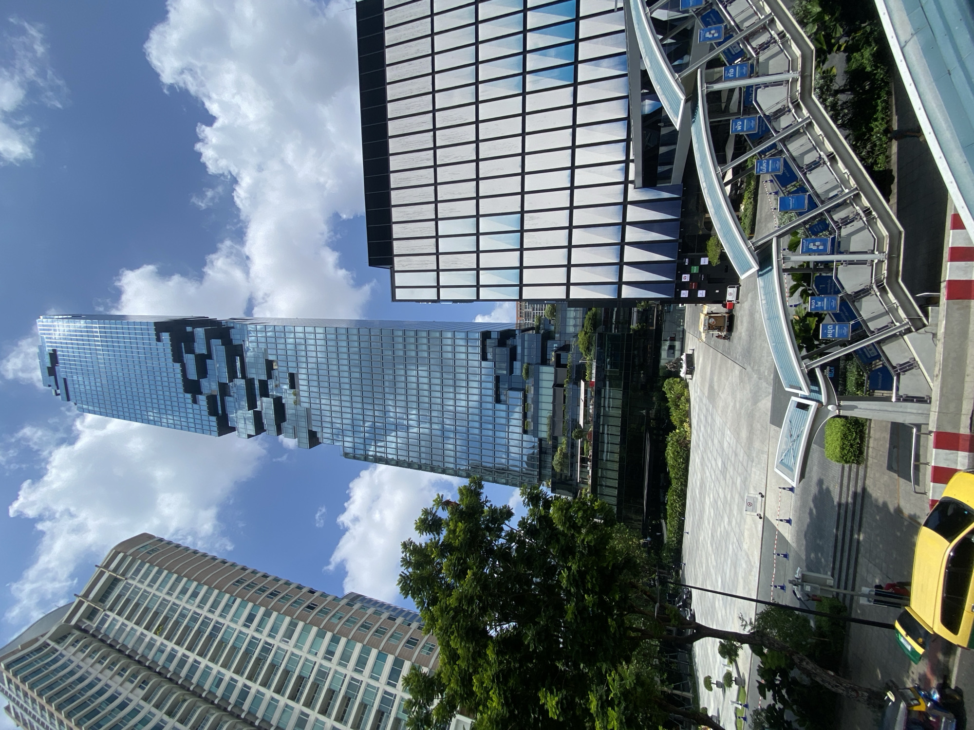 The image depicts an urban scene in Bangkok, Thailand, featuring several tall buildings under a partly cloudy sky. The most prominent building is the King Power MahaNakhon, a skyscraper with a distinctive pixelated or fragmented design. To the left of the MahaNakhon is a curved, tan-colored building with many windows. Adjacent to the MahaNakhon is another modern office building with a grid-like facade. In the foreground, there is a pedestrian skywalk with blue signage. Below the skywalk, a paved area leads to the buildings. A green tree partially obstructs the view of the buildings at the center of the scene. A yellow taxi is partially visible on the right side of the image. The photo appears to have been taken from an elevated position, possibly from another building or a skywalk.