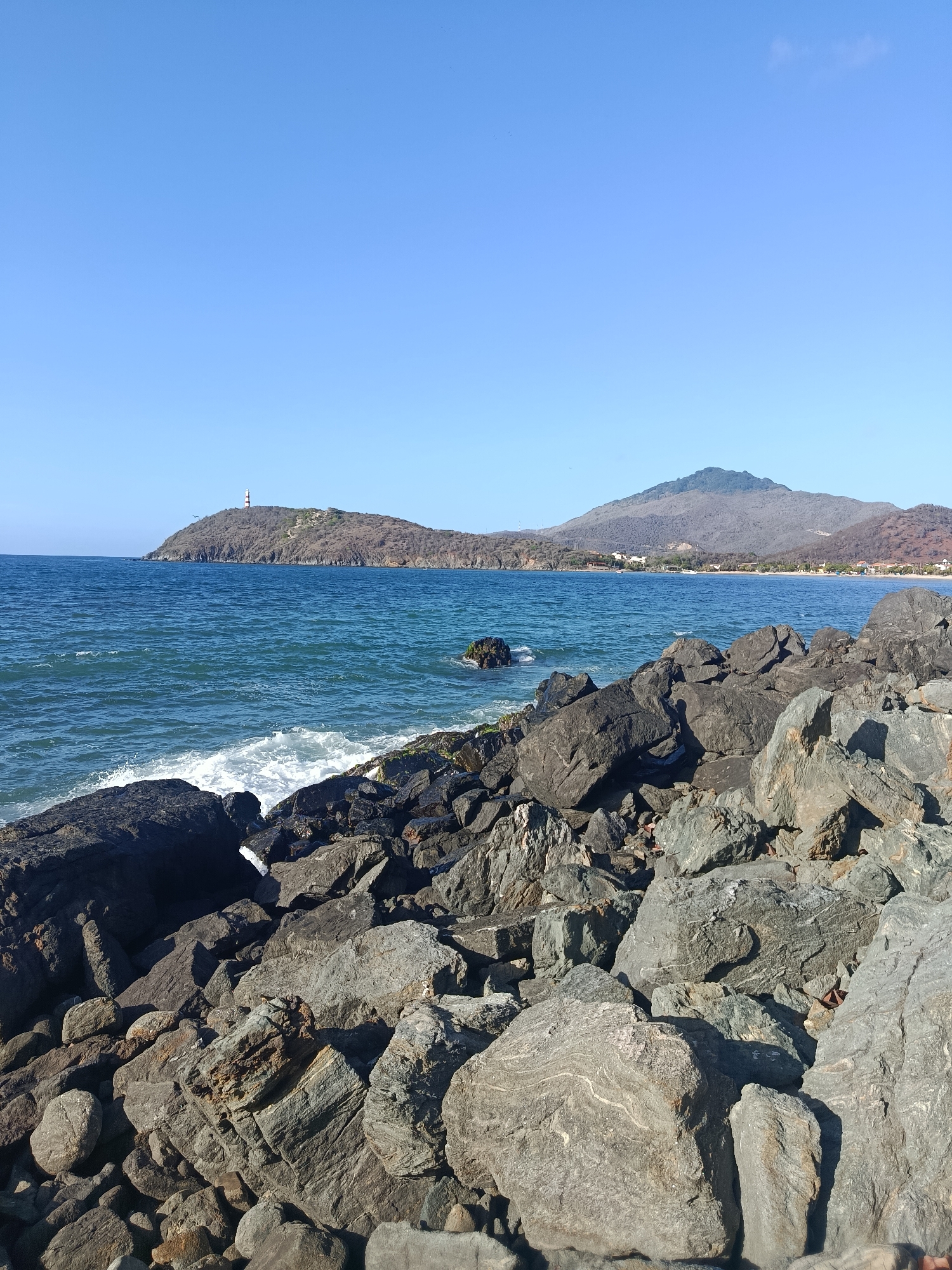 The image depicts a coastal scene in Pedro González, Venezuela. The foreground is dominated by a rocky shoreline, with large, gray boulders of varying sizes scattered across the frame. The rocks show natural textures and some appear to have lighter-colored veins.
Beyond the rocks, the vibrant blue ocean stretches out, with visible waves creating white foam as they break against the shore. In the middle ground, a distant, hilly peninsula rises from the sea, topped with a lighthouse that has a black and white striped design. The land on the peninsula appears arid and covered with sparse vegetation.
Further in the distance, a row of buildings, presumably part of the town of Pedro González, is visible along the coastline. Behind the town, larger, more rounded mountains form the backdrop, with their slopes displaying shades of gray and brown. The sky above is a clear, bright blue, indicating fair weather. There are no people or animals visible in the image.