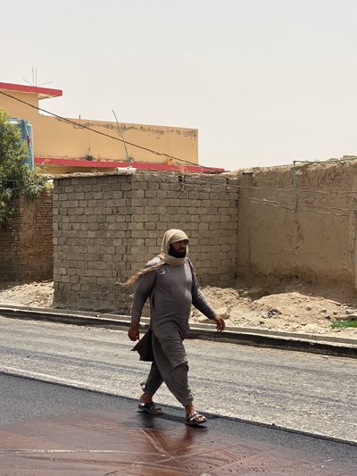 A man is walking on a newly paved asphalt street in an urban area of Kandahar, Afghanistan. The man is wearing a traditional grey salwar kameez, sandals, and a head covering draped around his neck. Behind him are buildings constructed from brick and stucco, with one building featuring a red trim. The street surface has a wet sheen, and the scene is illuminated by bright, overcast daylight.