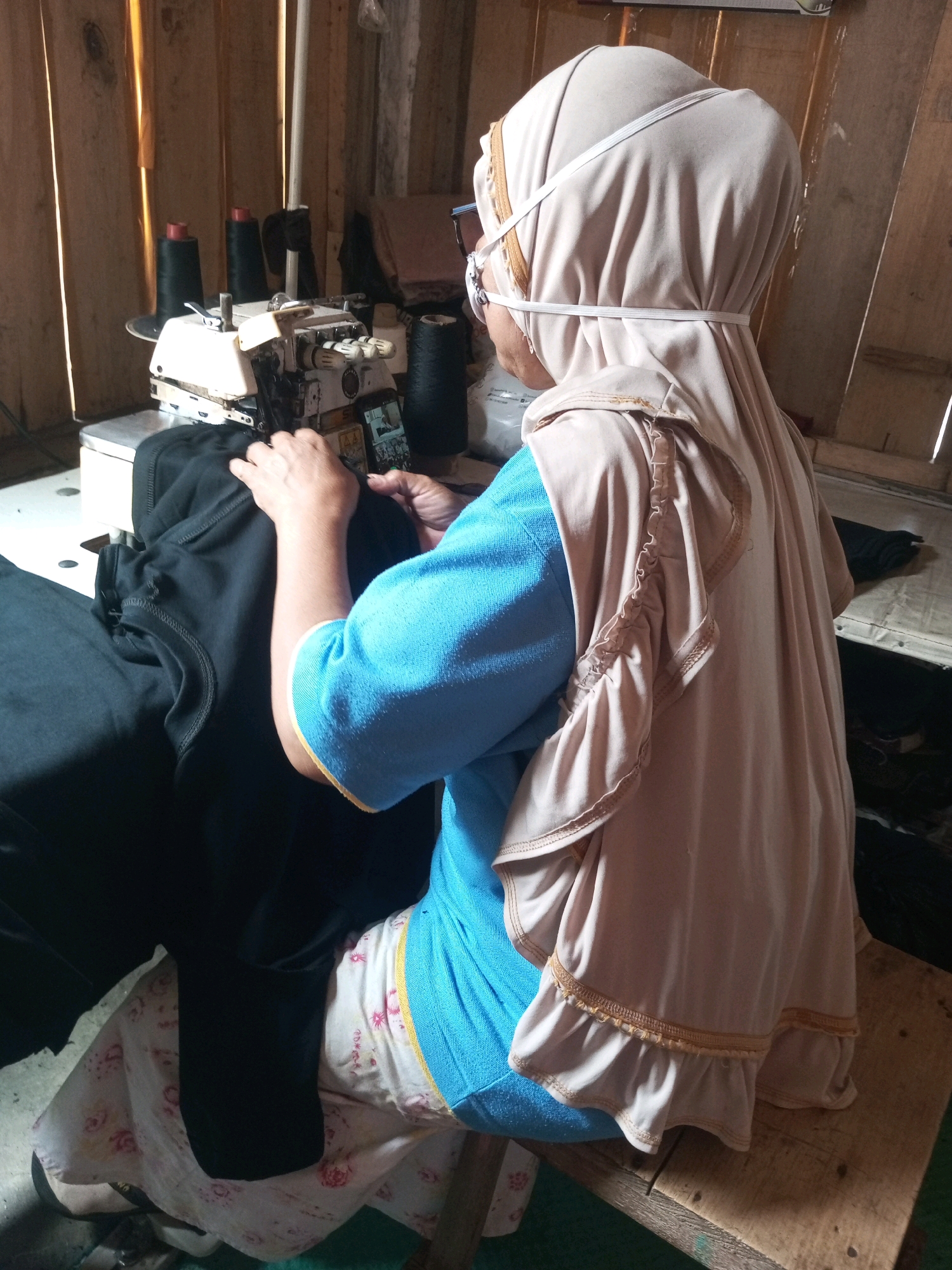 An overhead shot captures a person in a hijab and blue shirt seated at a sewing machine, working on a black garment. The environment appears to be a workshop in Kalibeber, Indonesia, with wooden walls and spools of thread on the machine. The person's hands are actively engaged with the fabric and the machine.