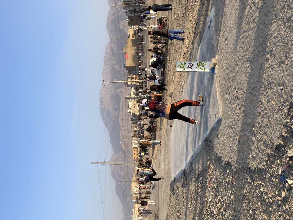 The image displays an informal outdoor cricket match in a dusty, unpaved field within Quetta City Tehsil, Pakistan, under a clear, light blue sky. In the foreground, a male batsman, wearing a multicolor patterned long-sleeved shirt, black pants with orange text, and a white baseball cap, is positioned on a concrete pitch, holding a cricket bat in a striking stance. A dark-colored cricket ball is visible near him. Opposite the batsman, a male bowler, dressed in a dark long-sleeved shirt and pants, is captured mid-action, having just released the ball, with his left leg elevated. Adjacent to the batsman's end of the concrete pitch, a white rectangular sign with green Urdu text is partially visible.
The mid-ground is densely populated with several dozen male spectators, observing the game. Many are standing, while others are seated on motorcycles and at least one white car parked around the perimeter of the playing area. Low-rise buildings with flat roofs are visible behind the spectators, indicating a populated urban or semi-urban setting. Overhead power lines crisscross the scene, supported by metal utility poles.
The background features large, rugged, and arid mountains under the clear sky. The ground throughout the playing area is uneven, consisting of loose earth, small rocks, and scattered debris. The scene is illuminated by natural light, casting discernible shadows, suggesting either morning or late afternoon.