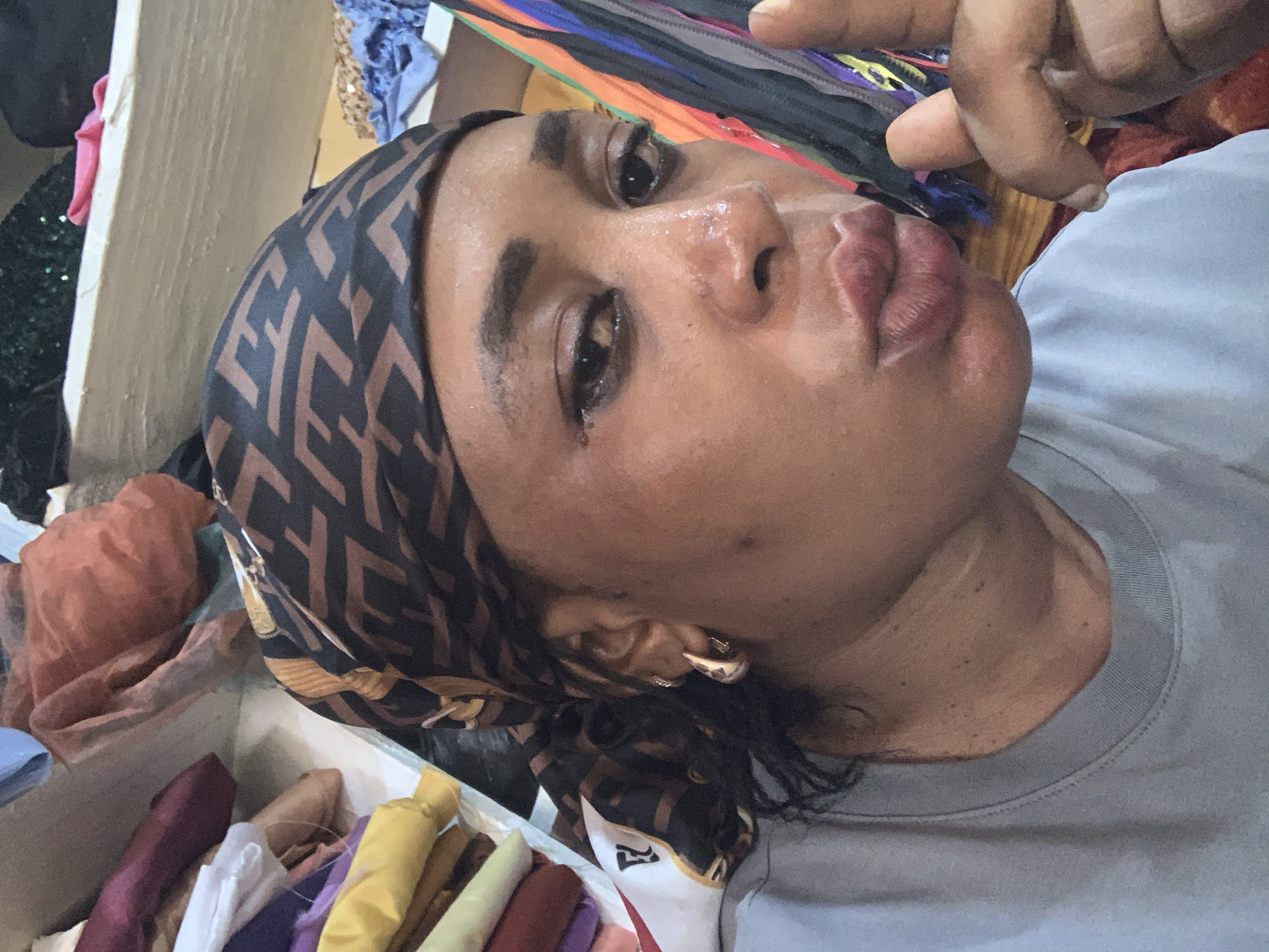 A close-up, eye-level selfie depicts an adult female individual with a dark complexion, positioned in an indoor commercial setting. She is wearing a light grey crew-neck t-shirt and a black and brown patterned headwrap featuring a repeating "F" logo, indicative of the Fendi brand. Her dark, curly hair is visible beneath the headwrap on her left side. Her face exhibits a shiny sheen, possibly from sweat or oil, and her full lips are pursed in a pouting expression. Her eyes are lined with dark makeup and gaze directly at the camera. A gold-toned hoop earring is visible in her left ear. Her right hand is raised, with the index finger slightly curled upward.
The background suggests a fabric or textile shop, likely located in Sabon Tasha, Nigeria. To the left, stacks of various rolled or folded fabrics are visible, including yellow, light green, maroon, and white textiles, some on shelves. A white fabric with a red "F" logo (Fendi) is also present. To the right, multiple vertical strips of multi-colored fabric, possibly ribbons or samples, hang. The upper background shows additional shelves containing crumpled and folded fabrics of different colors, including an orange-brown and a green sequined material. The environment appears functional and densely packed with merchandise.