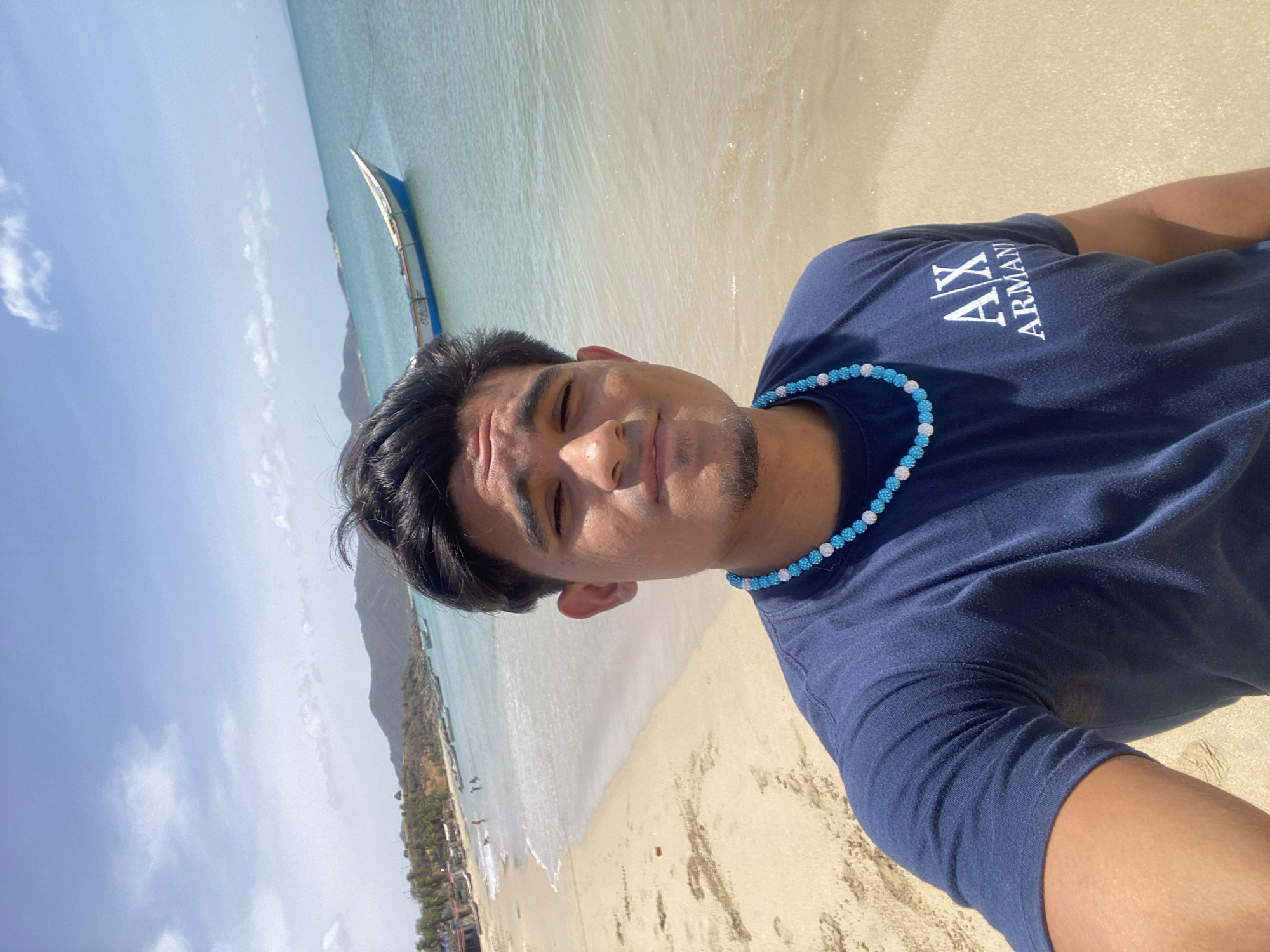 A medium-shot, eye-level, self-portrait depicts a man on a beach. He wears a navy blue t-shirt with white "AX ARMANI" text on the left side of his chest and a light blue beaded necklace. The beach setting features light beige sand, clear shallow water, and a distant boat. In the background, there is a coastline with buildings and a mountainous landscape. The sky is partly cloudy with a light blue hue. This scene is located in Juan Griego, Venezuela.