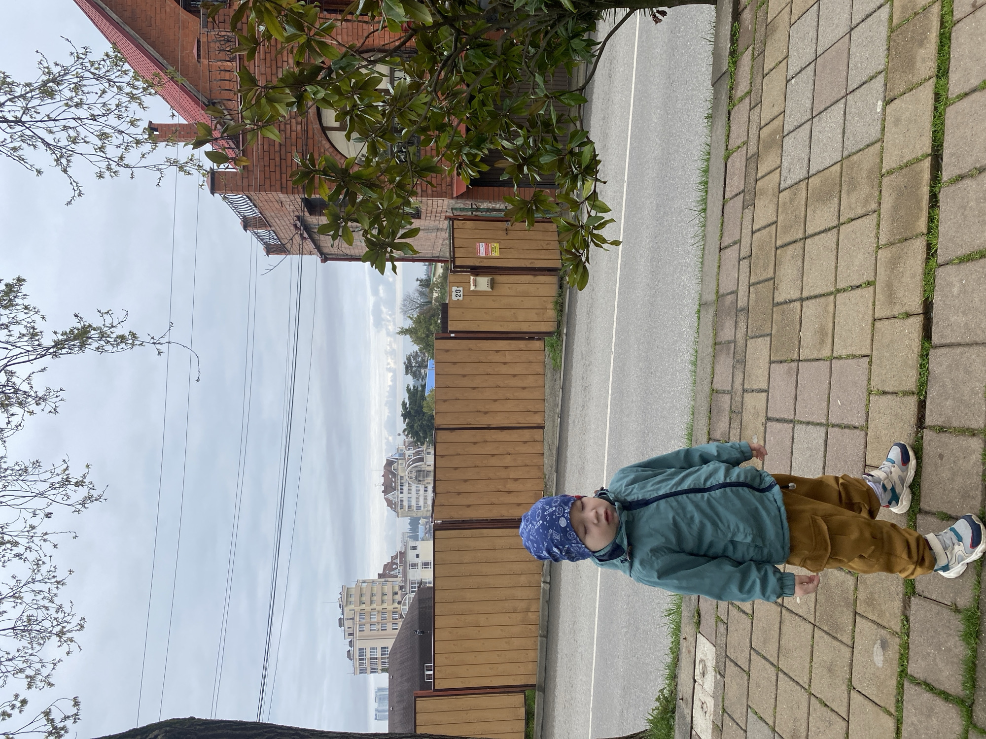 The image captures an outdoor urban scene in Sochi, Russia, under an overcast sky. A young child stands on a square-tiled concrete sidewalk in the foreground, positioned slightly left of the frame's vertical center. The child wears a blue patterned beanie, a teal zippered jacket, brown cargo pants, and multi-colored athletic shoes; their eyes appear closed or squinted.
To the child's right, a paved asphalt road with a white solid line runs parallel to the sidewalk. Directly behind the child, a tall wooden fence with vertical planks extends horizontally across the mid-ground. A metal mailbox and the house number "28" are visible on a section of this fence, along with a red and white warning sign.
In the mid-ground right, partially obscured by a tree with large dark green leaves, stands a multi-story brick building featuring a red tiled roof, a prominent chimney, and a balcony with a black wrought-iron railing. The building exhibits arched windows. In the background, beyond the wooden fence, several other multi-story residential buildings with varying architectural styles are visible. Overhead, multiple utility lines traverse the light grey sky, which is partially framed at the top by tree branches with small, light green leaves. A tree trunk is visible on the far left edge of the frame.