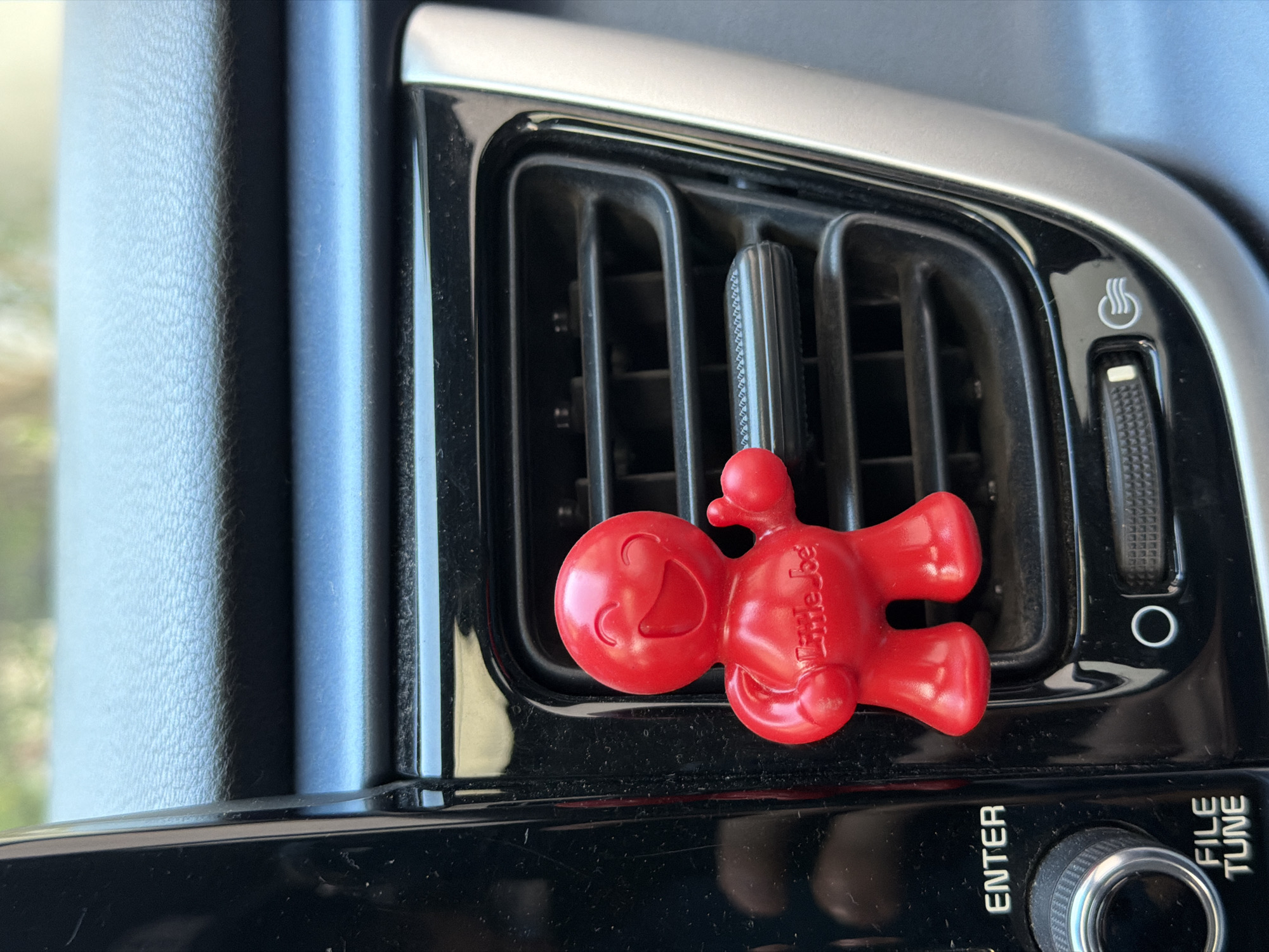 A close-up shot inside a car shows a red "Little Joe" air freshener clipped to a black air vent. The vent is situated within a silver trim and is positioned above the "ENTER" and "FILE TUNE" buttons and knob. The image was not taken in the city of Cuautla, Mexico.