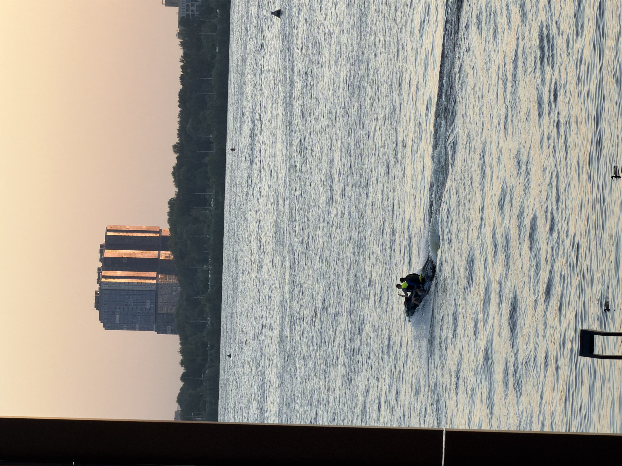 The image depicts a scene on the Kazanka River in Kazan, Russia. The photo appears to have been taken from an elevated position, possibly from a building overlooking the river. Two individuals are riding a jet ski on the water. The river is slightly choppy with visible water ripples. The skyline includes a modern high-rise building with a glass facade reflecting sunlight and a line of trees along the riverbank. The sky is a light, warm color, suggesting either sunrise or sunset. The lower part of the picture contains some darker structures from the building that appear to be part of a balcony.