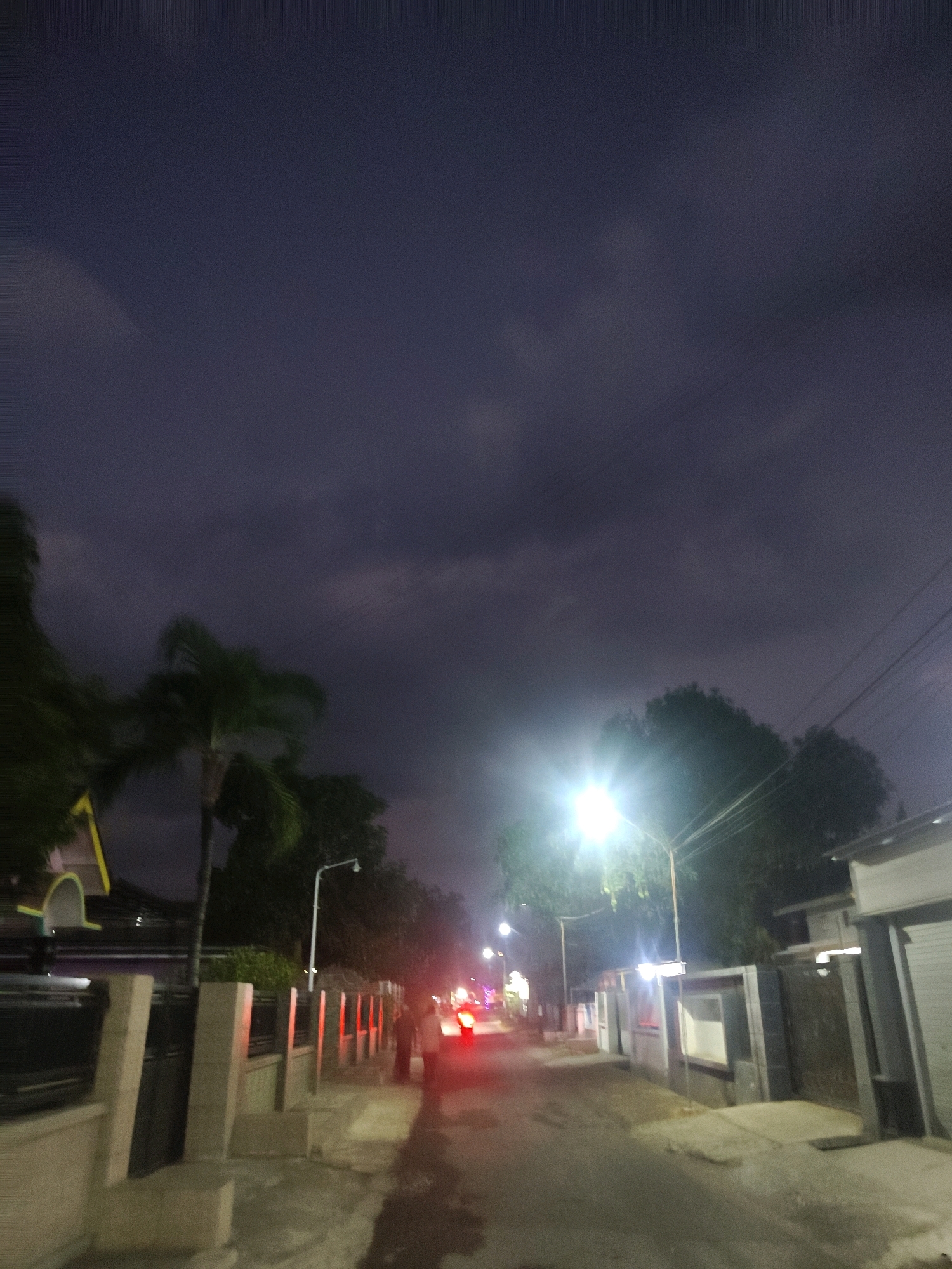 The image captures a street scene at dusk or night in Cermee, Indonesia. The sky is a dark, cloudy purple. A street lamp illuminates the left side of the road, casting a bright light on trees and surrounding buildings. A palm tree is visible on the left side of the street, and several houses with fences line the right side. A person, blurred by motion, is walking on the sidewalk. Red taillights of a vehicle are visible in the distance, suggesting traffic. The overall atmosphere is dimly lit, with streetlights and vehicle lights providing the primary illumination.