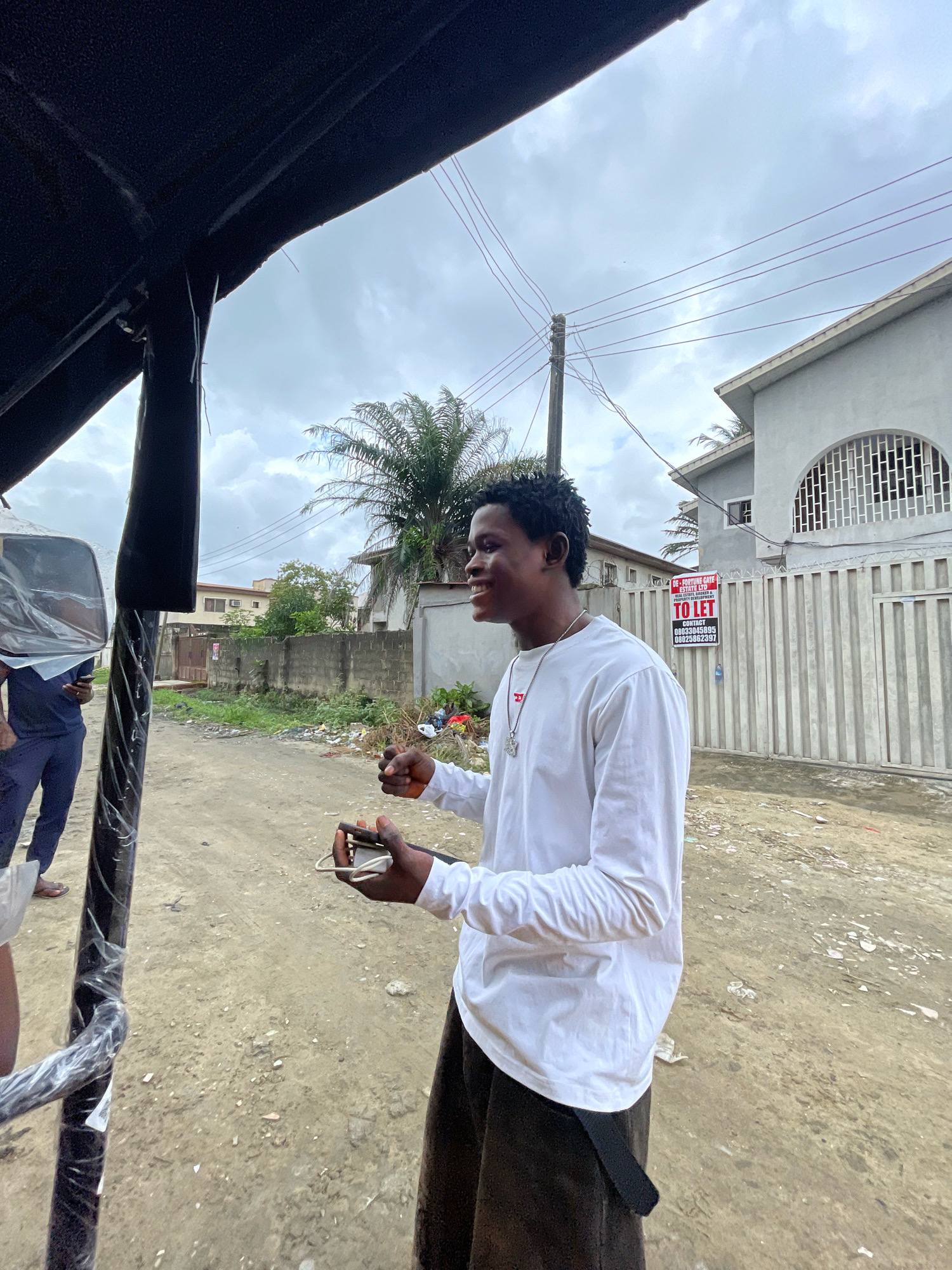 In Ado, Nigeria, a young Black man wearing a white long-sleeved shirt and dark pants stands in front of a grey building with a white fence, holding a cell phone and what appears to be cash. He is smiling and looking to his left. The man is standing next to a dark, possibly black, structure that is partially visible and covered in plastic wrap. In the background, there are other buildings, a palm tree, a utility pole with wires, and a "TO LET" sign on the fence. The ground is dirt and debris. Another person, wearing a blue shirt and dark pants, is visible to the left of the frame. The sky is overcast.