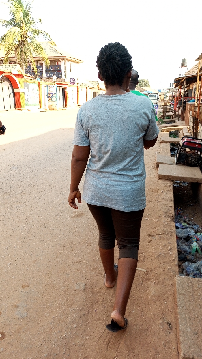 An outdoor street scene in Sabon Tasha, Nigeria, is depicted from behind a walking individual. The primary subject, an adult female, faces away from the camera, wearing a light grey short-sleeved t-shirt, dark brown capri-length leggings, and dark sandals. Her hair is styled in dark dreadlocks. She walks on a reddish-brown dirt road with scattered small stones. To her immediate right, a section of an open concrete drainage ditch, filled with plastic bags and refuse, parallels the road. A red and black portable generator rests on the concrete lip of this ditch. Beyond the ditch, a line of single-story structures, possibly market stalls or shops with various facades and corrugated roofs, extends into the distance. To the left, further down the road, a multi-story building with a light-colored facade, decorative blue patterns, and a dark roof is prominent, flanked by a tall palm tree bearing coconuts. Smaller businesses with distinct signage, including a purple sign reading "CANTONE," are visible at street level below the larger building. Another person wearing a green shirt is partially visible to the right of the primary subject, also moving away. The sky is bright and clear.