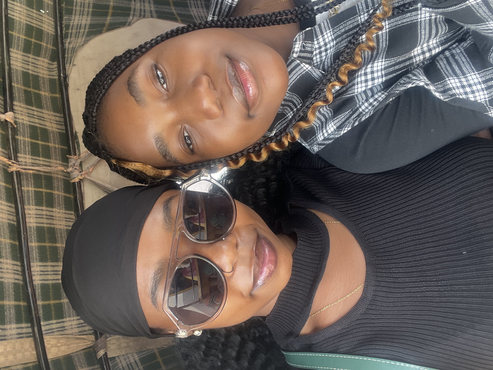 Two young Black women are taking a selfie in what appears to be an open-air vehicle, possibly a rickshaw, with a checkered fabric canopy overhead. The woman on the left is wearing a black headscarf, large clear-rimmed sunglasses, and a black ribbed top with a cut-out detail at the neck, adorned with a thin gold chain. Her hair is dark and curly. The woman on the right has her hair in black braids with some lighter brown highlights, and she is wearing a black and white checkered shirt. Both women are looking at the camera and smiling. The background shows the interior of the vehicle, including the patterned canopy and possibly some upholstery. There are no visible elements in the image that specifically indicate the location as Barakin Lerawi, Nigeria.