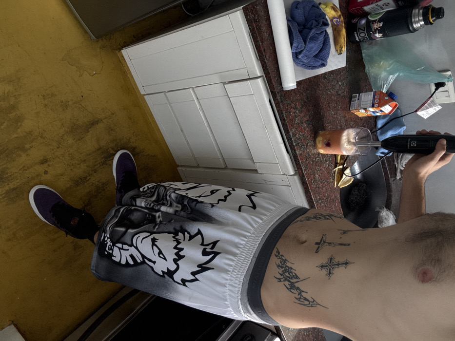 An overhead view depicts a shirtless individual operating an Electrolux immersion blender in a kitchen located within Buenos Aires, Argentina. The person's torso, visible from mid-chest to feet, exhibits dark body hair and several black ink tattoos on the left side, including an inverted cross, another cross-like symbol, and abstract designs. They are dressed in white, grey, and black patterned shorts featuring cartoonish characters, and purple sneakers with white soles.
The individual's right hand holds the black immersion blender, immersing it into a clear glass on a dark red granite countertop. The glass contains a frothy, reddish-pink liquid with yellow fruit pieces, indicative of a smoothie in preparation. The blender's power cord extends towards a white wall outlet.
The countertop also displays two ripe bananas on a white sheet, a folded blue fabric, a small orange juice carton (Baggio Naranja), and a black thermos bottle with "ZOR" branding. A dark circular sink basin, containing a dark scrubber and a banana peel, is visible behind the blending area. A light blue plastic bag hangs from the wall above the sink. White, painted wooden base cabinets with multiple drawer fronts are situated beneath the countertop. Portions of a silver refrigerator are visible on the left and a dark appliance (likely a stove) on the right. The floor is a worn, yellow-painted surface with visible stains and scuffs.