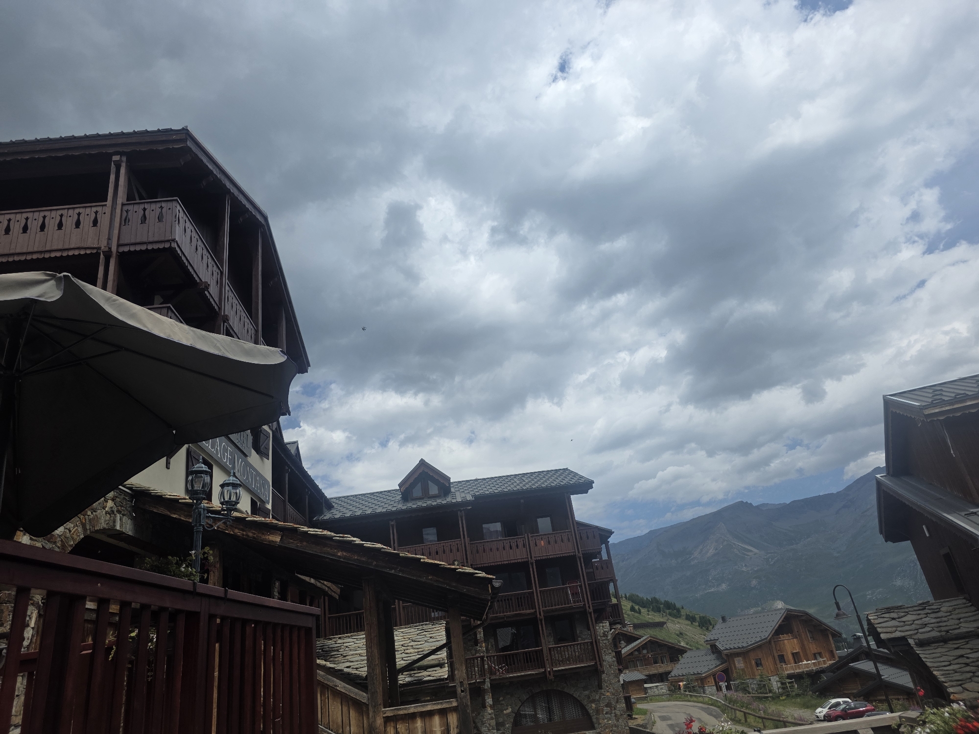 In Tignes, France, a view captures the architectural style of a mountain village with a cloudy sky. Dark brown, multi-story buildings with wooden balconies and stone foundations dominate the foreground. One building, to the left, features a dark umbrella partially obscuring its balconies and a sign that reads "VILLAGE MONTANA." Further to the right, more wooden chalets with dark roofs are nestled on a hillside, with a winding road and a few parked cars visible. In the background, a range of majestic, hazy mountains stretches across the horizon. The atmosphere is overcast, with dramatic cloud formations filling the sky.