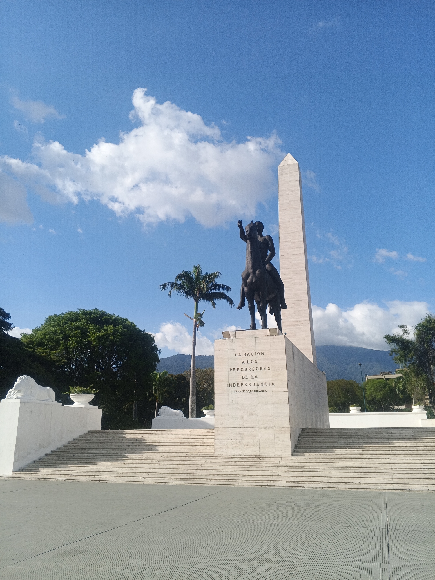 The image depicts a statue of a man on horseback atop a stone pedestal with an obelisk in Caracas, Venezuela. The statue and monument are centrally located in the frame, with a wide staircase leading up to the pedestal. Inscribed on the pedestal is the phrase "La Nacion a los Precursores de la Independencia, Francisco de Miranda." To the left of the monument, a lush green tree and a palm tree are visible. Other architectural features, including what appear to be lion statues, are to the left and right of the staircase. In the background, a mountain range can be seen under a blue sky with scattered white clouds. The foreground is a gray, paved plaza.