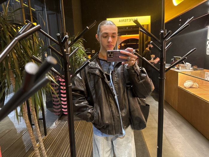 An individual with pulled-back hair, wearing a dark leather bomber jacket over a light blue garment and grey sweatpants, is depicted taking a selfie in a reflective surface. The individual's right hand holds a smartphone with an American flag-themed case, displaying multiple rings on the fingers. A black, multi-branched coat rack is prominent in the foreground. The scene is an interior of a cafeteria in Yerevan, Armenia. To the left, a large potted plant is visible. In the background, behind the coat rack, a male with a beard, wearing a dark shirt and beanie, stands behind a wooden service counter, holding a reddish cup. The counter features a glass display case containing several light-colored baked goods or pastries. The word "CAFETERIA" is visible in reverse, illuminated in yellow above the counter. The walls are dark, and the floor is a light grey. Artificial lighting is the primary light source.