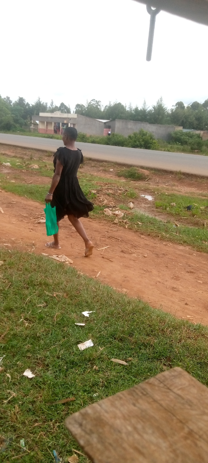 A woman wearing a black dress and sandals walks on a dirt road while carrying a green plastic bag. The road is flanked by green grass and a gray concrete wall, a small structure is visible in the background to the left of the scene. The road has a paved surface. The woman is walking from right to left. The woman is in focus, the background is out of focus. The scene takes place in a rural area of Kivaywa, Kenya. The road is lined with trees on the right side. The scene is overcast and grey. There is a wooden object and debris in the foreground. There are a few small puddles on the road. The scene is taken from a low angle, looking up at the woman. The image quality is poor.