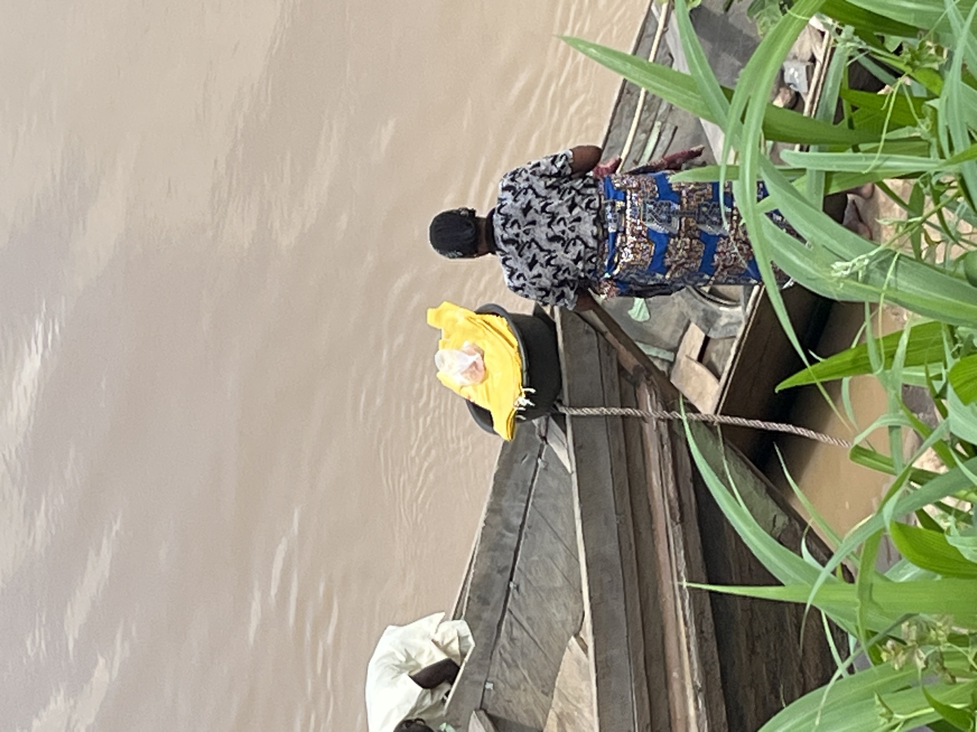 The image portrays a tranquil river scene in Idah, Nigeria, featuring two individuals in traditional wooden boats amidst lush green vegetation. The calm river water, a muted brown or beige, occupies the left side of the frame.
In the foreground, a person is partially visible in a wooden boat, seen from behind, wearing a light-colored or white garment. Further back, another individual, also facing away from the camera, is situated in a second wooden boat. This person wears a black and white patterned short-sleeved top and a vibrant blue fabric wrap or skirt adorned with gold and brown patterns, typical of traditional attire. Their head is covered by a dark cap, and they are in a bending or squatting posture, seemingly engaged in an activity within the boat.
A thick rope connects the two boats, suggesting they are moored together. A bright yellow object, possibly a bag or piece of clothing, rests near the bow of the front boat. The right side of the scene is framed by abundant, tall green foliage, likely reeds or grasses, characteristic of a riverside environment. The natural daylight suggests it is daytime. No text is visible in the image. The scene captures a quiet moment of daily life on the river.