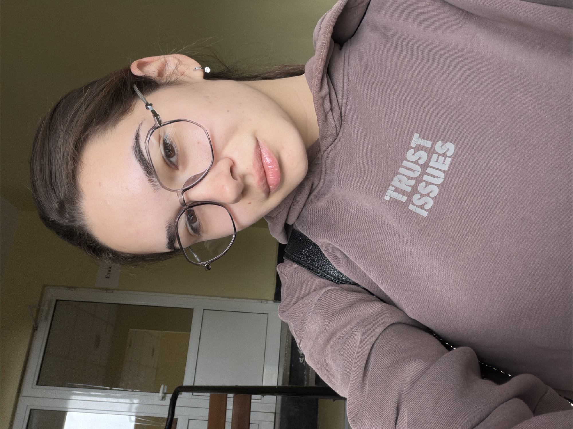 A young woman with dark hair is depicted in a selfie-style photo. She is wearing glasses and a brown hoodie with the words "TRUST ISSUES" printed on it. She has a hoop earring in her left ear. The background includes a white window with a black frame. The image may have been taken indoors, possibly in Sofia, Bulgaria, as implied from the request for location information. Further environmental and relational details are not readily available.