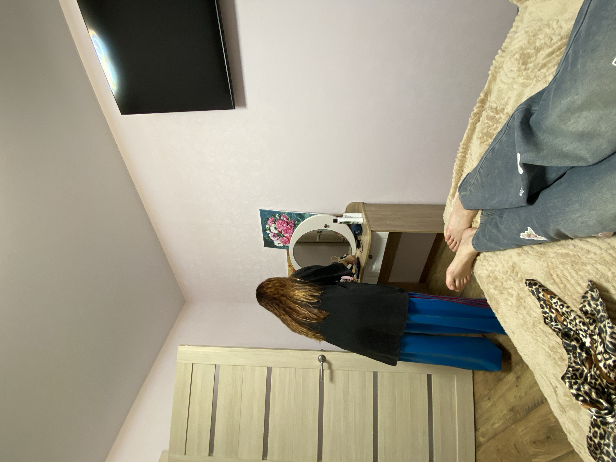 The image depicts an indoor scene, likely a bedroom, with a person standing at a vanity table. The person has long, wavy brown hair and is wearing a dark top and bright blue pants. They appear to be interacting with items on the vanity, which includes a mirror and various cosmetic products. To the left of the vanity is a light-colored wooden door. In the foreground, on a textured beige surface, are a pair of legs wearing denim jeans and a leopard-print fabric item. A television is mounted on the wall in the upper right portion of the image. The room has pale pink walls and a light-colored ceiling. The scene is consistent with a domestic setting in Votkinsk, Russia.