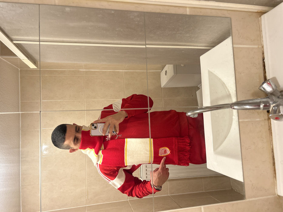 A person in a red Adidas tracksuit and a red and white scarf with a logo is reflected in a bathroom mirror. The person holds a phone, presumably taking the picture. A white sink with a silver faucet is to the right. Beige tiles cover the wall. The mirror shows a toilet and a partial view of the bathroom. The person points at the scarf's logo.